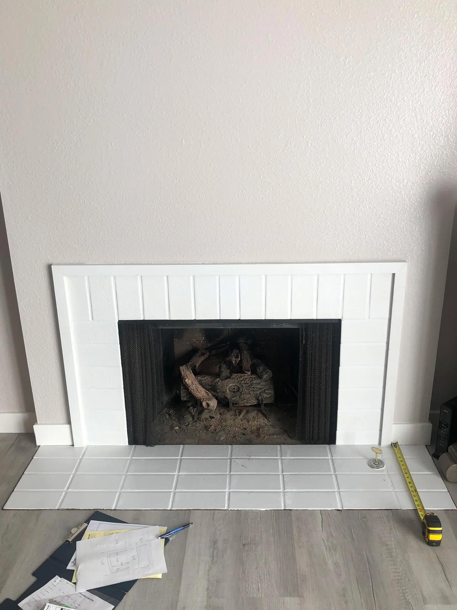 A white tiled fireplace with a black grate, surrounded by beige walls and a light wooden floor. A measuring tape and scattered papers are on the floor in front of the fireplace.