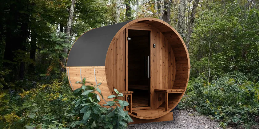 A cylindrical, wooden outdoor sauna with a black roof sits surrounded by dense, green foliage, and trees. The door is open, revealing an inviting interior with benches on either side.