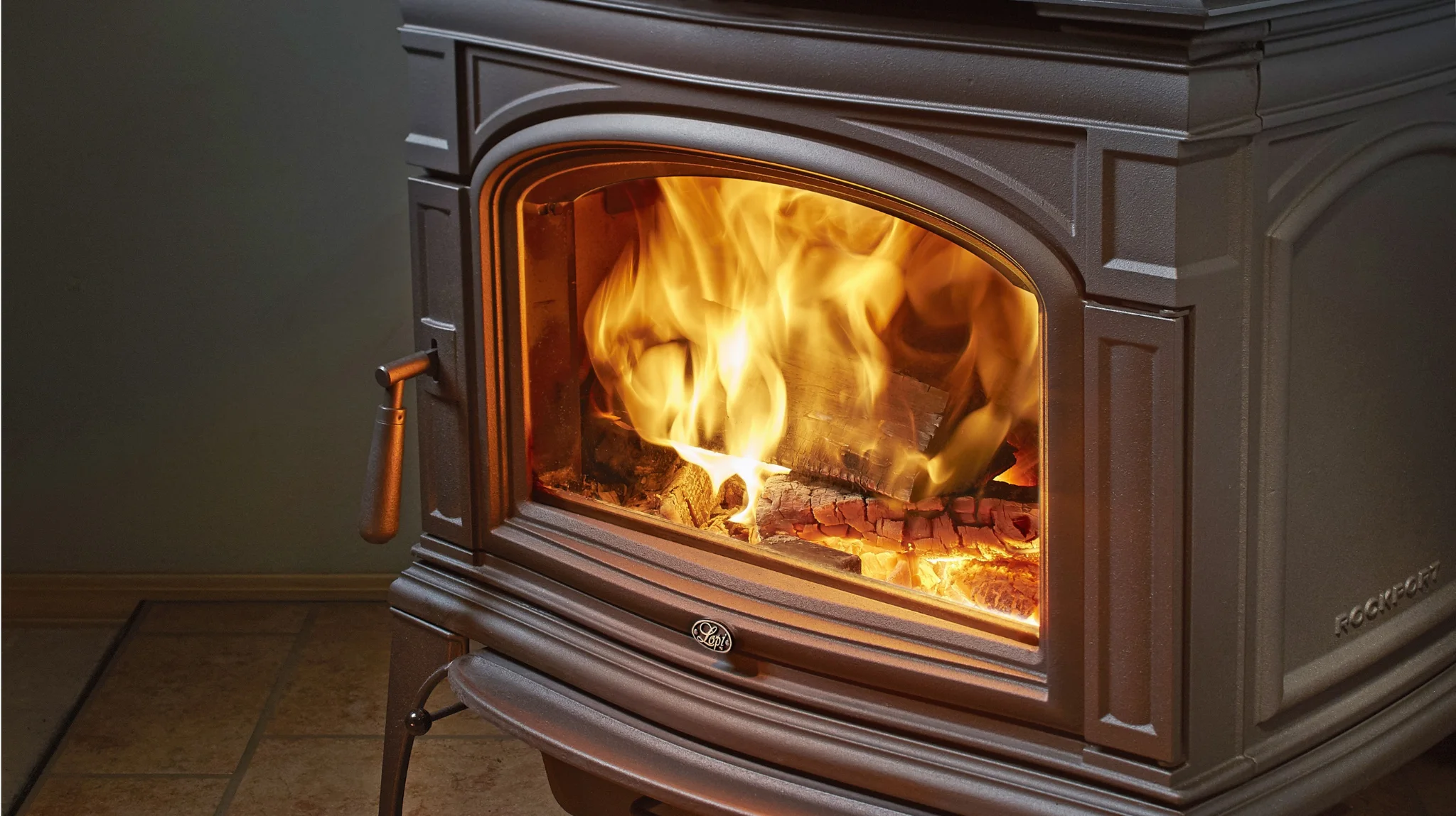A black wood-burning stove with a glass door showing a large, bright fire inside. The stove is indoors on a tiled floor with a handle visible on the left side.