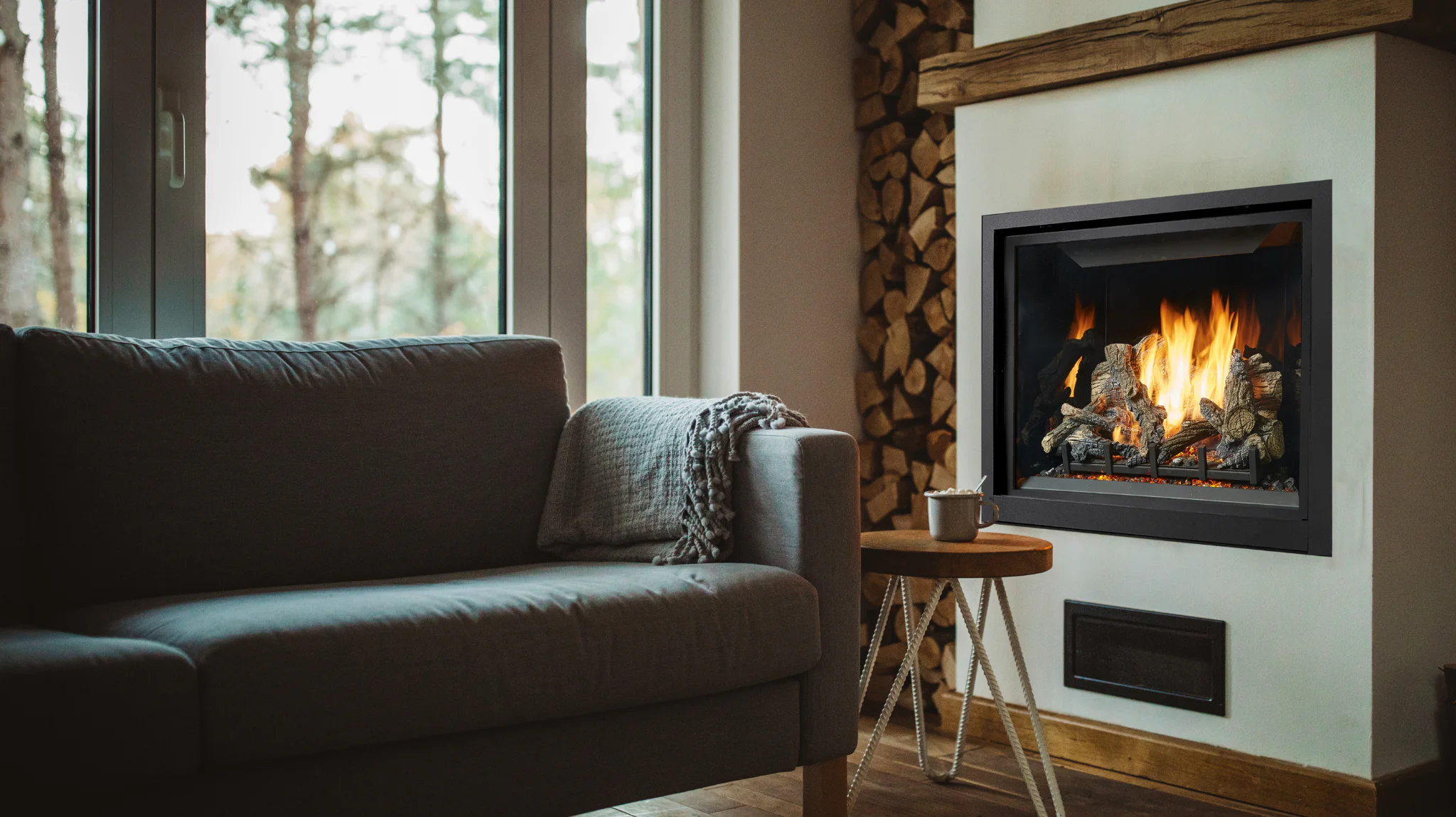 A cozy living room features a lit fireplace with stacked logs, a plush gray sofa adorned with a textured throw, and a small wooden side table with a mug on top.