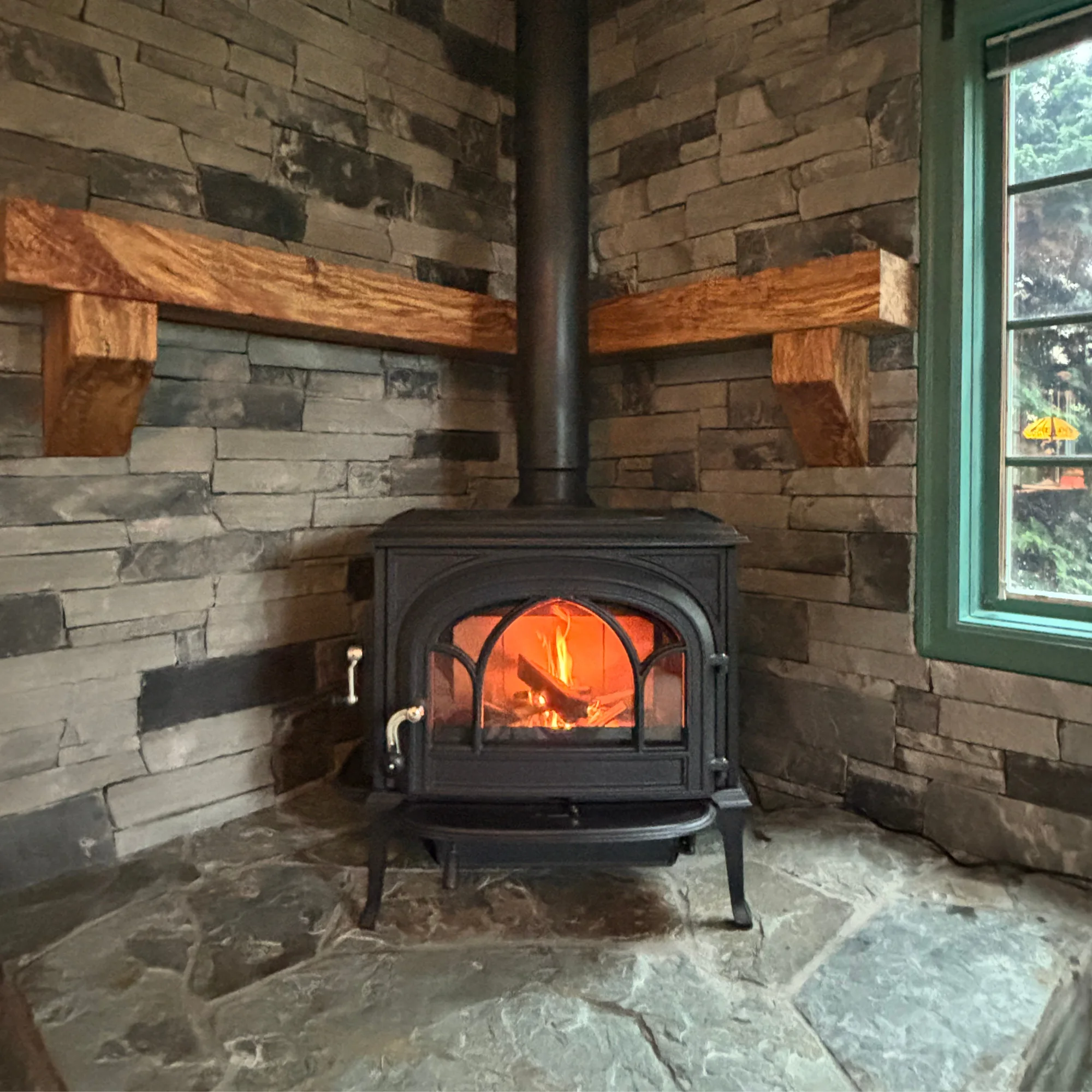 A black wood-burning stove with a fire inside sits on a stone floor against a stone brick wall with wooden shelves. A green-framed window is on the right side.