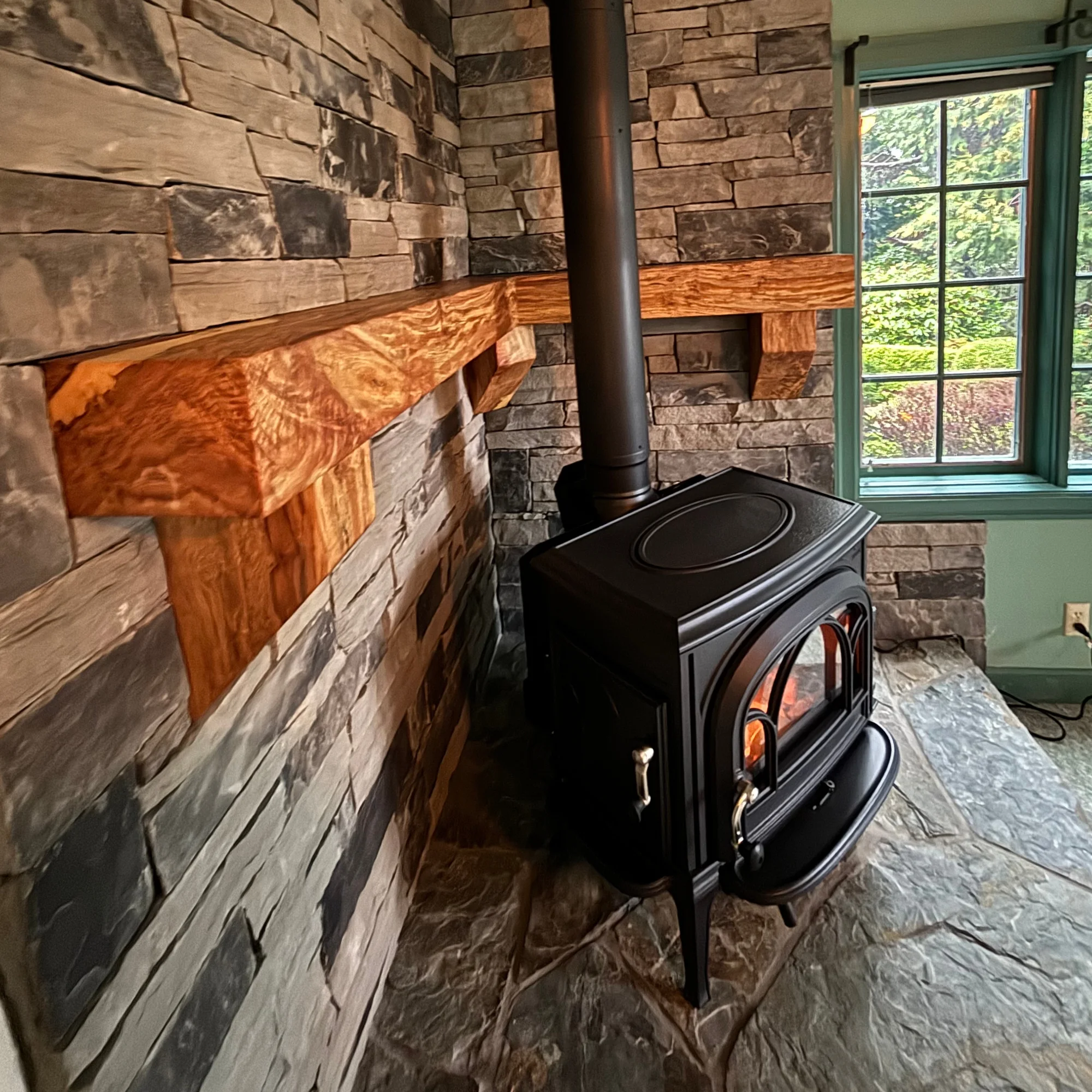 A black wood-burning stove with a visible flame, set against a stone wall with a wooden mantel. The room has a stone floor and a window with a green frame showing a garden outside.