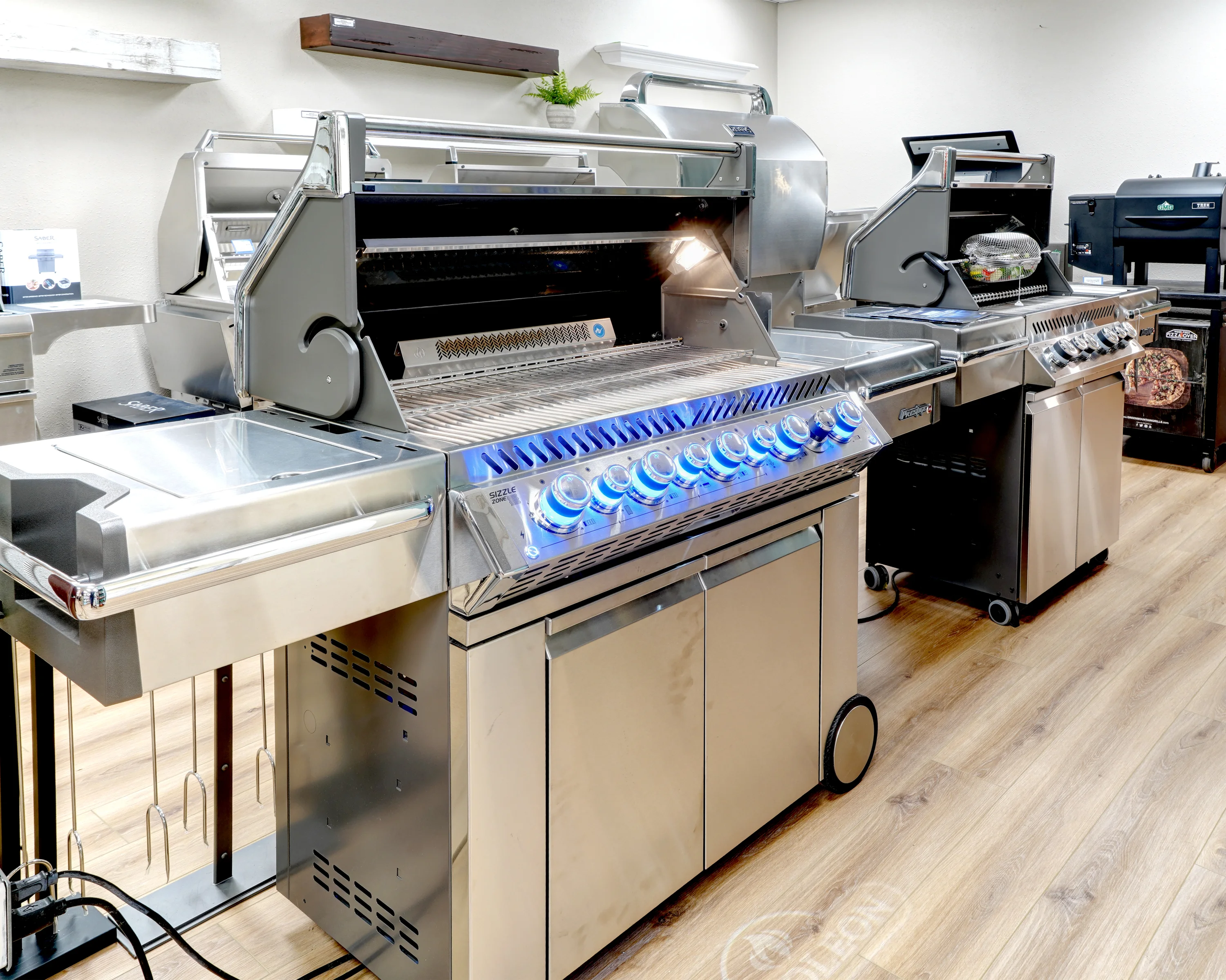 Showroom with three large stainless steel gas grills on display. Grills feature multiple burners, blue LED-lit dials, and various accessories. A wooden floor and white walls are visible.