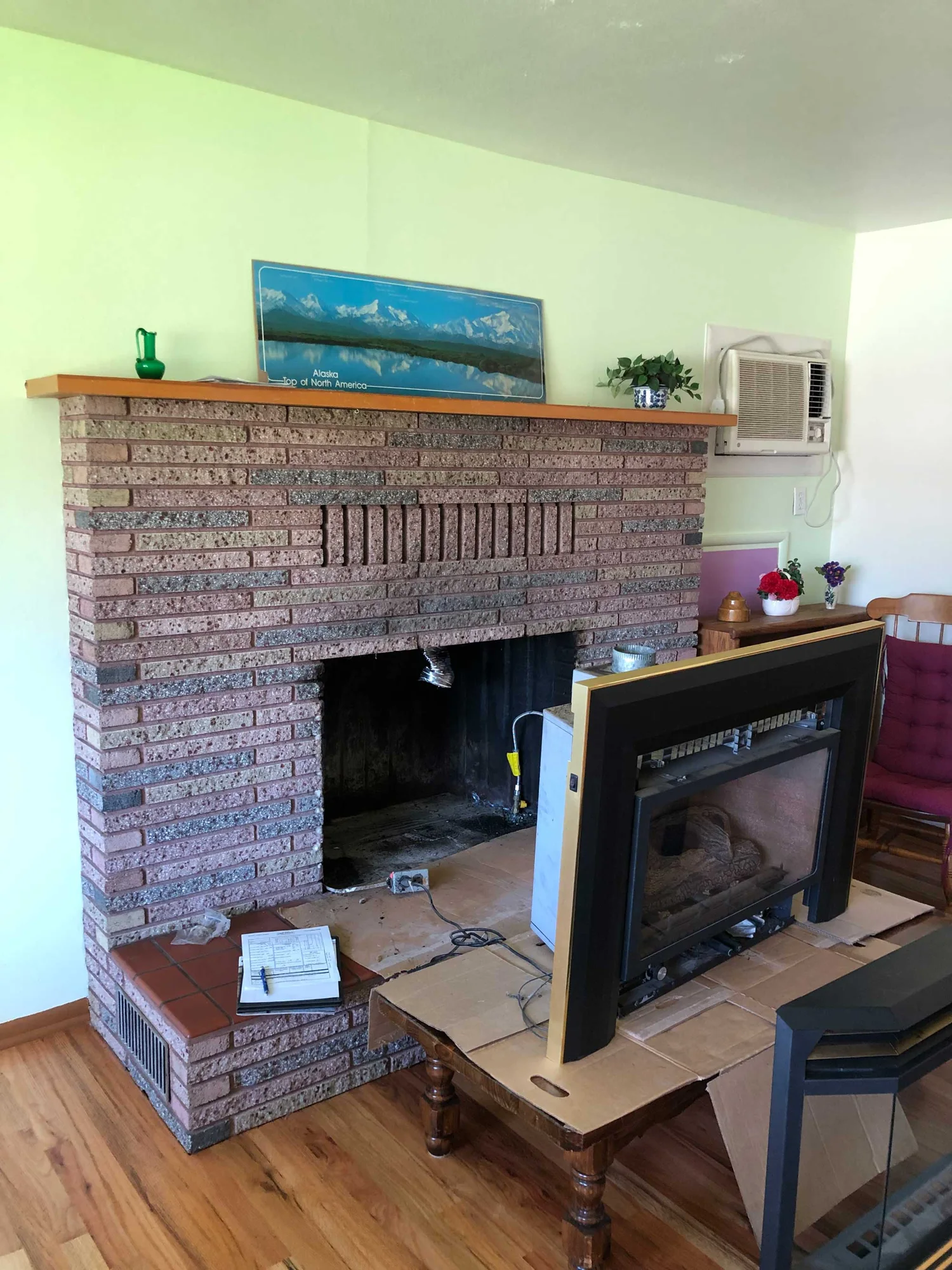 Brick living room fireplace with its metal insert removed and propped on a small table, mantel with landscape print and potted plants, window air conditioner