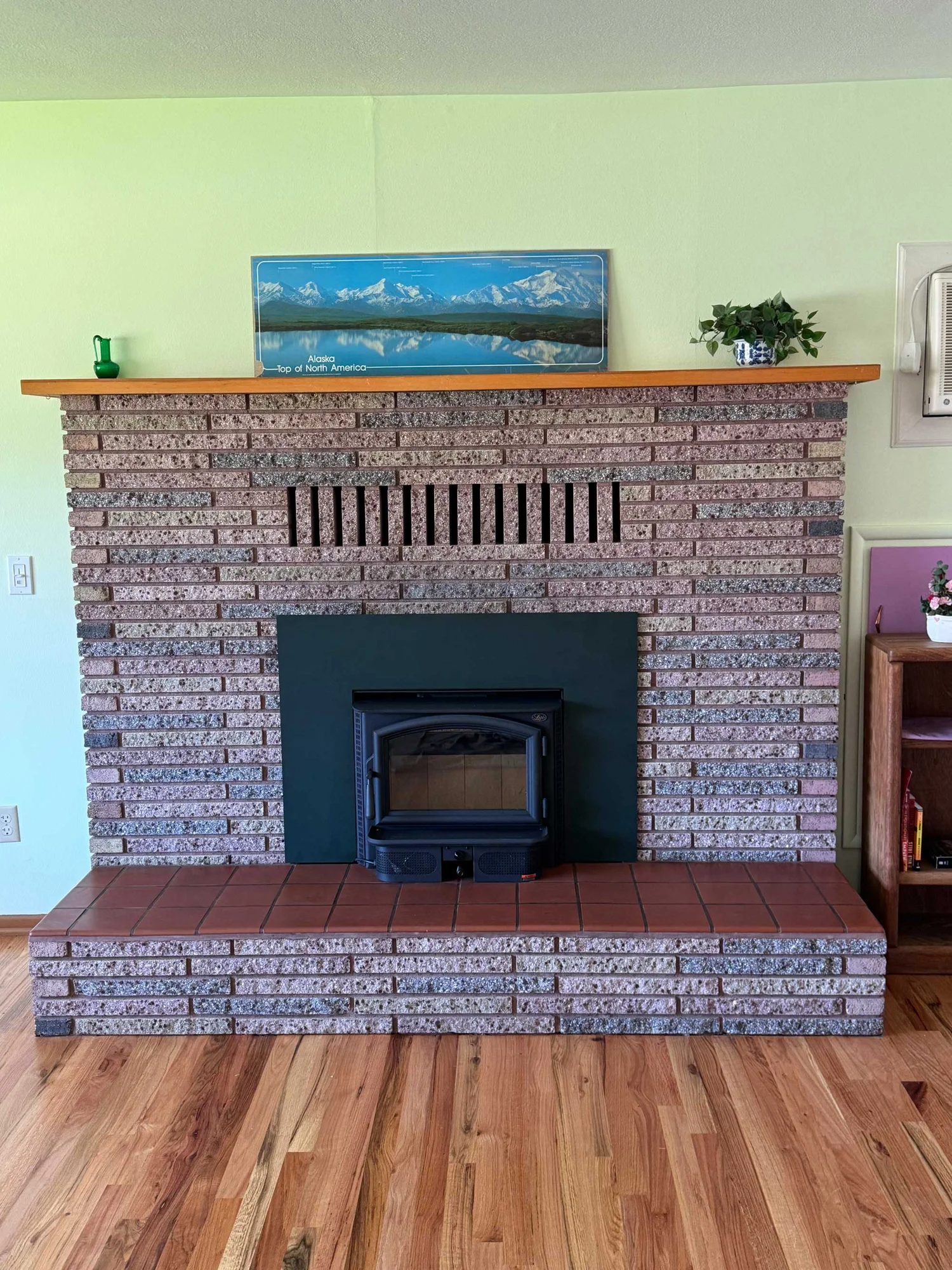Brick fireplace with black wood stove insert and tiled hearth, wooden mantel with Alaskan landscape print, small green pitcher and potted plant on pale green wall.