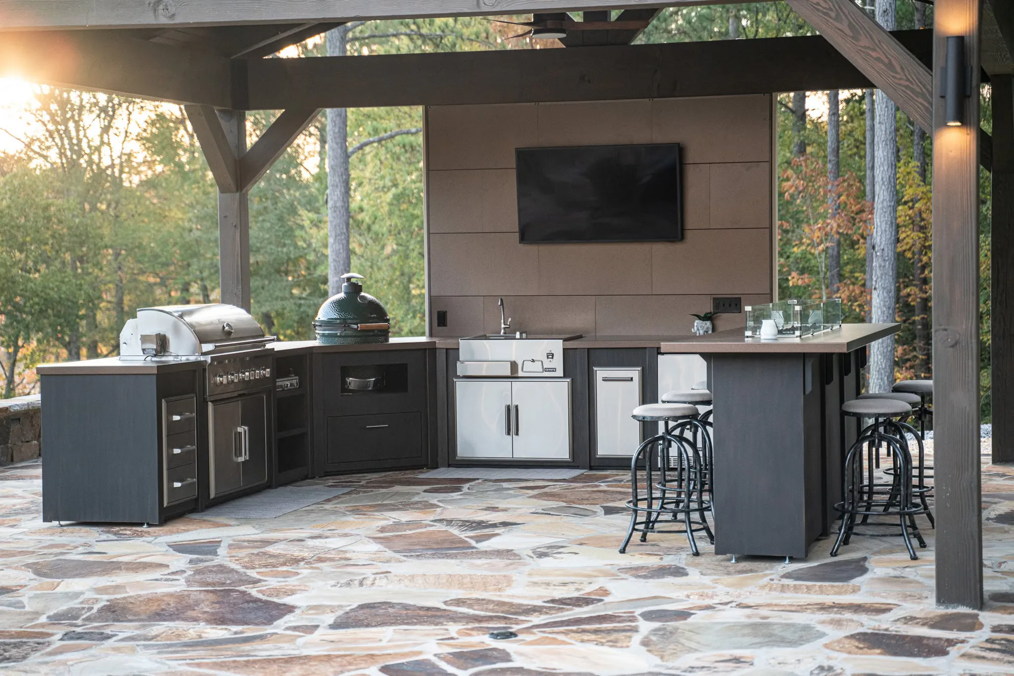 Covered outdoor kitchen with stainless grill, green kamado smoker, wall-mounted TV, bar counter and stools on a stone patio