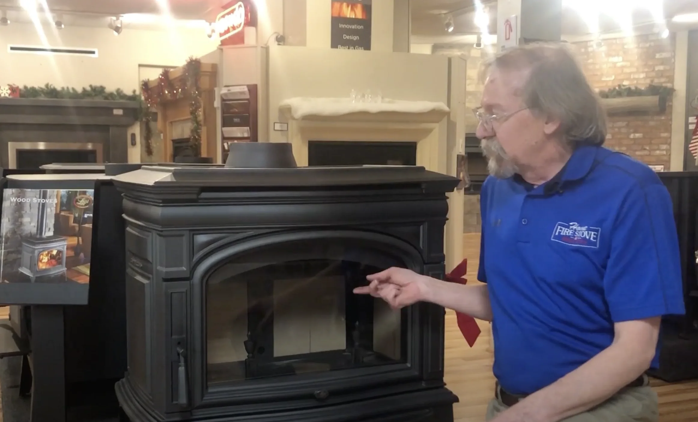 A man in a blue shirt is pointing at a black wood stove in a showroom with various fireplace models on display.