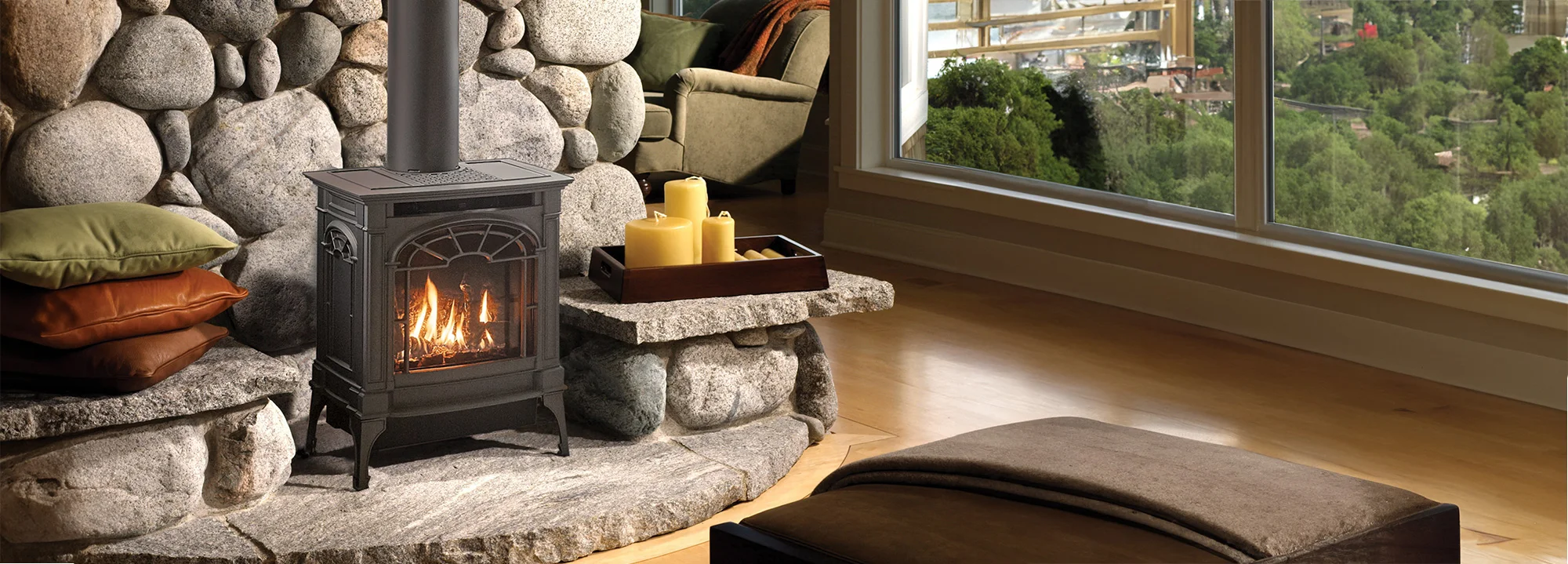 Cozy living room with a small black wood-burning stove on a stone hearth, with cushions on the left and candles on the right. Large windows show a scenic outdoor view.