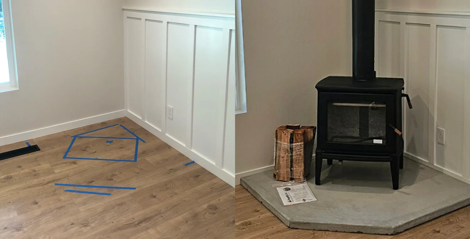 Corner room showing blue tape outline on floor marking stove location and a black wood stove installed on a raised concrete hearth with stacked firewood beside it