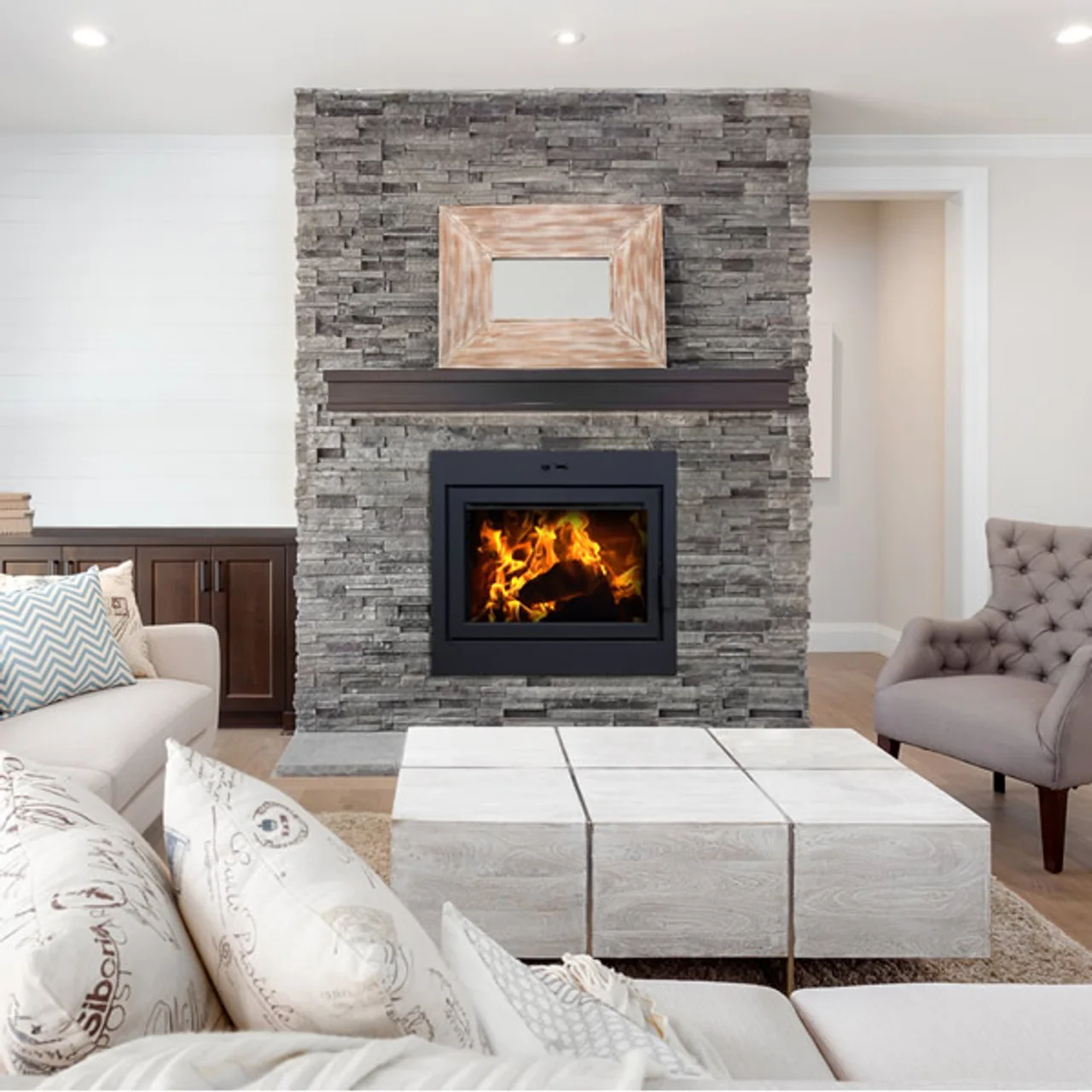 Modern living room with a stone fireplace, a wooden framed mirror above, and a lit fire. The seating includes a beige sofa and a gray tufted chair with decorative pillows and a wooden table.