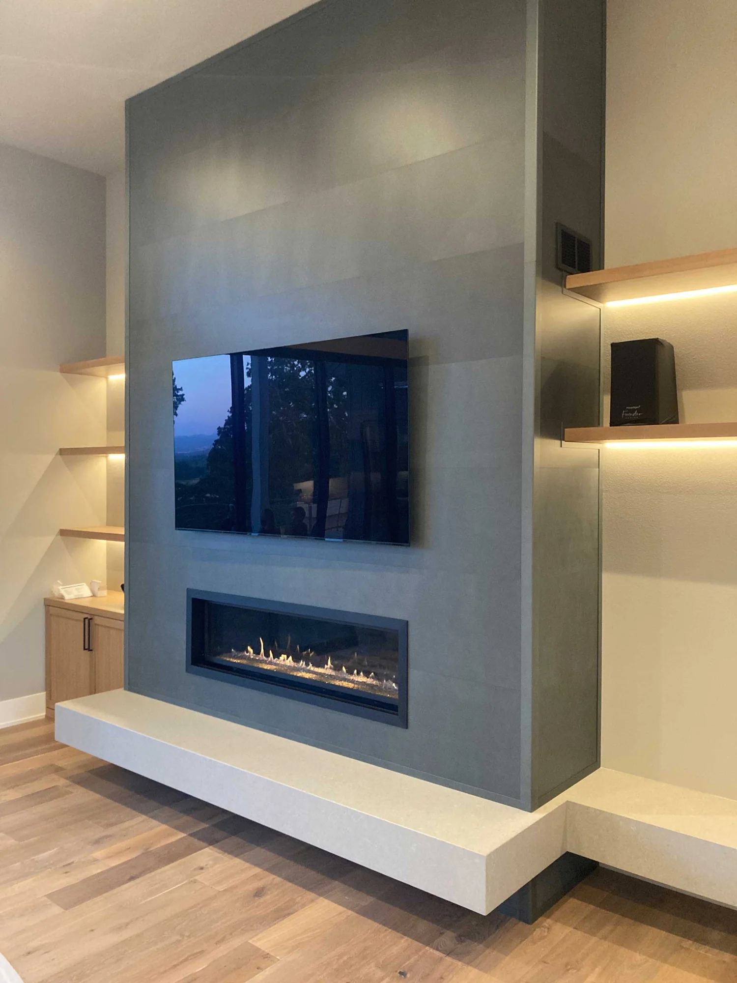 Modern living room wall with mounted flat-screen TV above a long linear fireplace and floating white hearth, wood floors and lit built-in shelves.