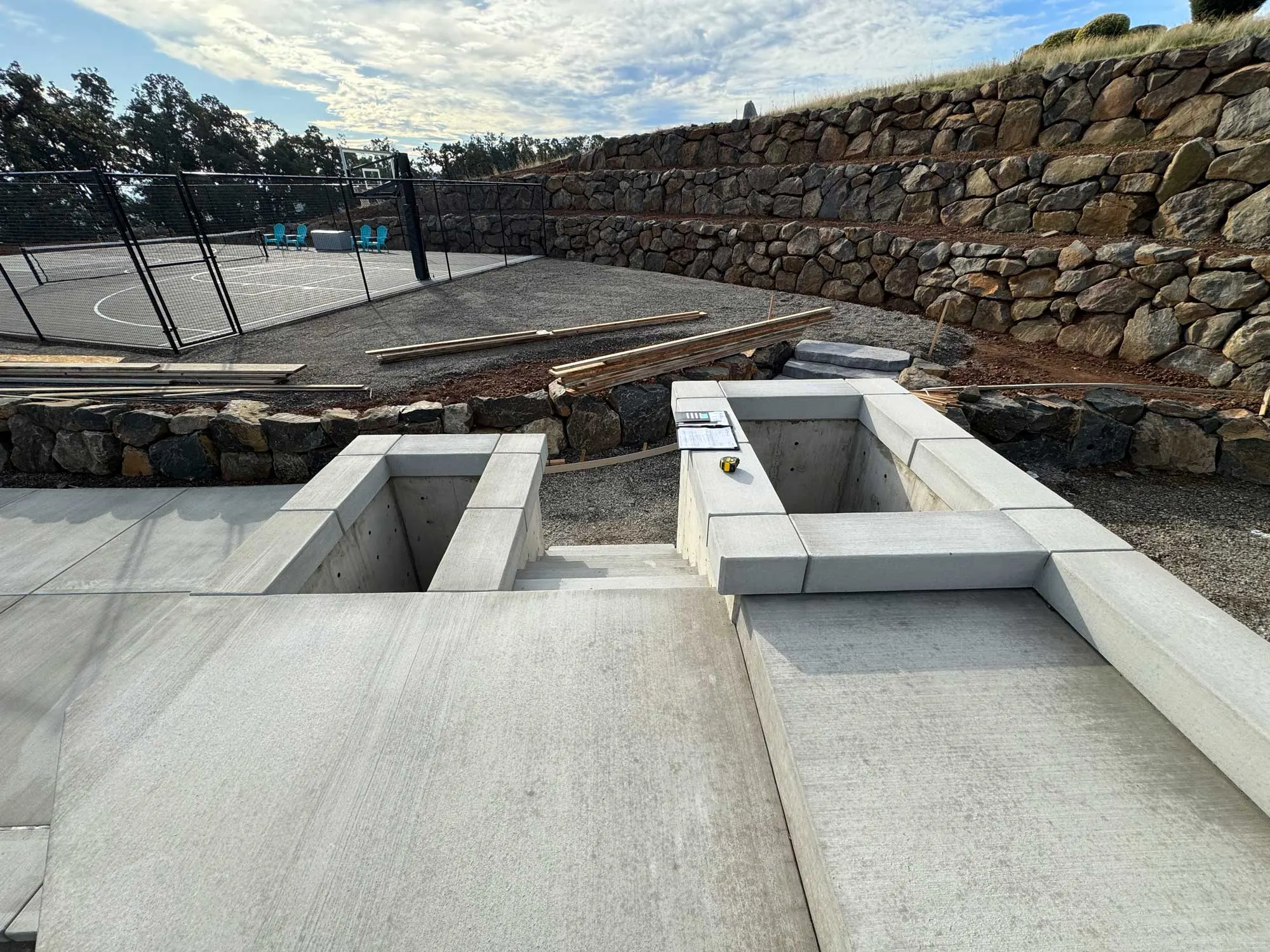 Concrete steps leading to a backyard with concrete planters, gravel pad, fenced basketball half court on left and terraced stone retaining walls on right