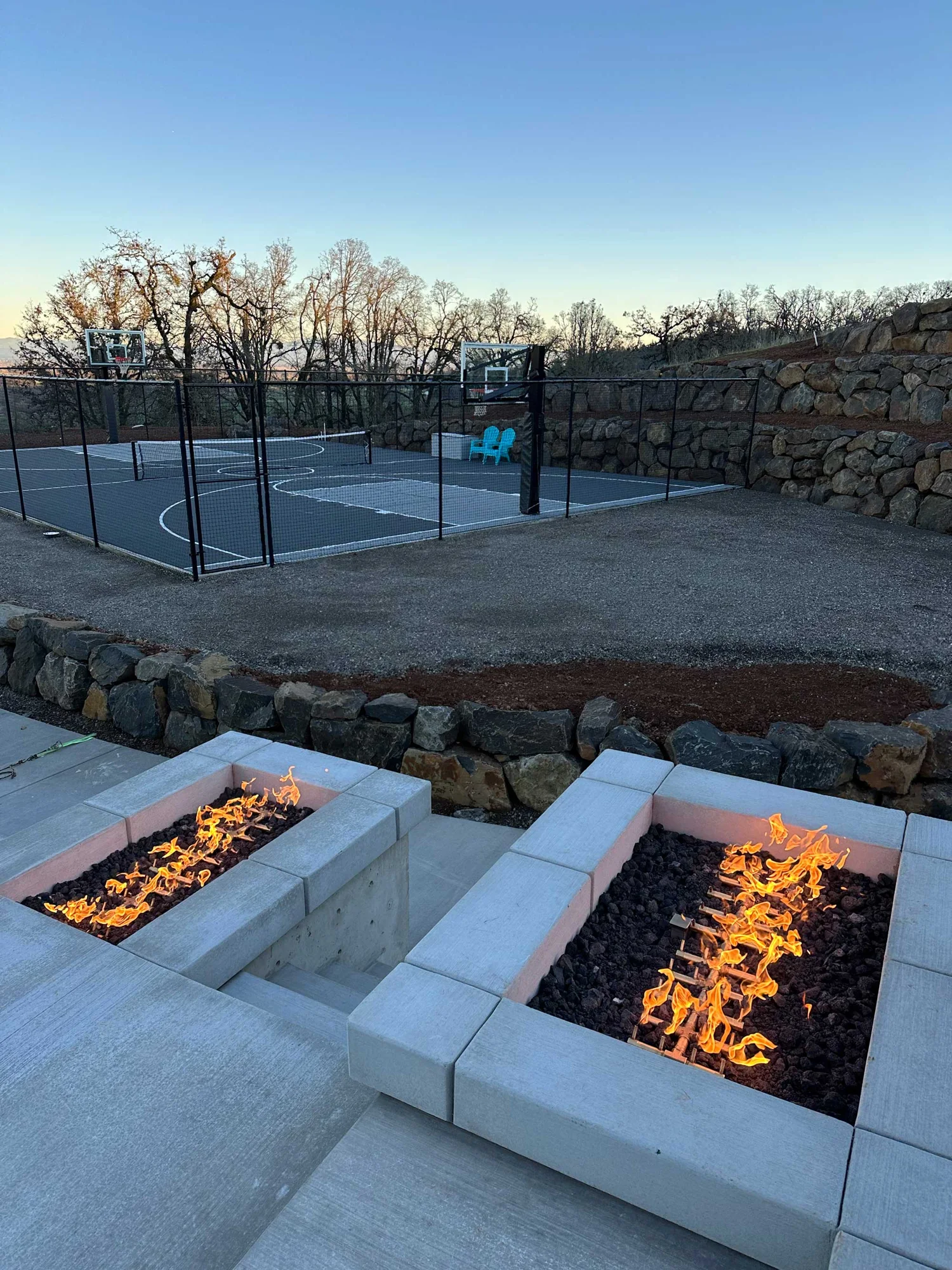 Two rectangular gas fire pits in concrete seating in foreground, fenced outdoor basketball court with turquoise chairs, stone retaining walls and leafless trees at dusk