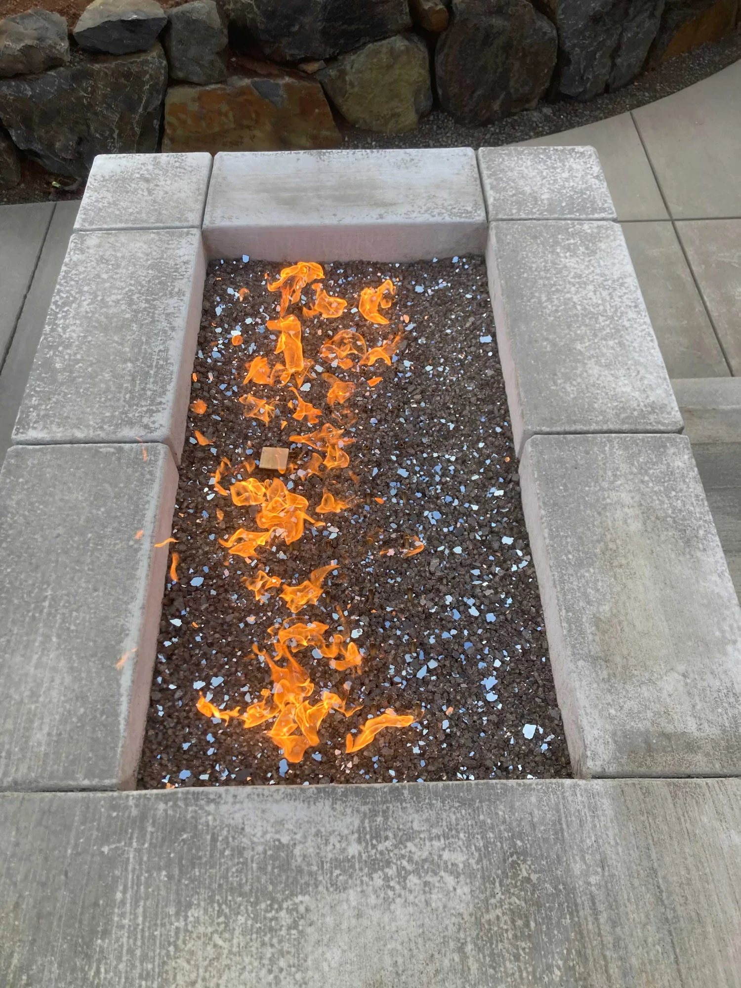 Rectangular outdoor gas fire pit with orange flames over dark lava rock and blue reflective glass pieces, surrounded by weathered concrete block seating and stone wall.