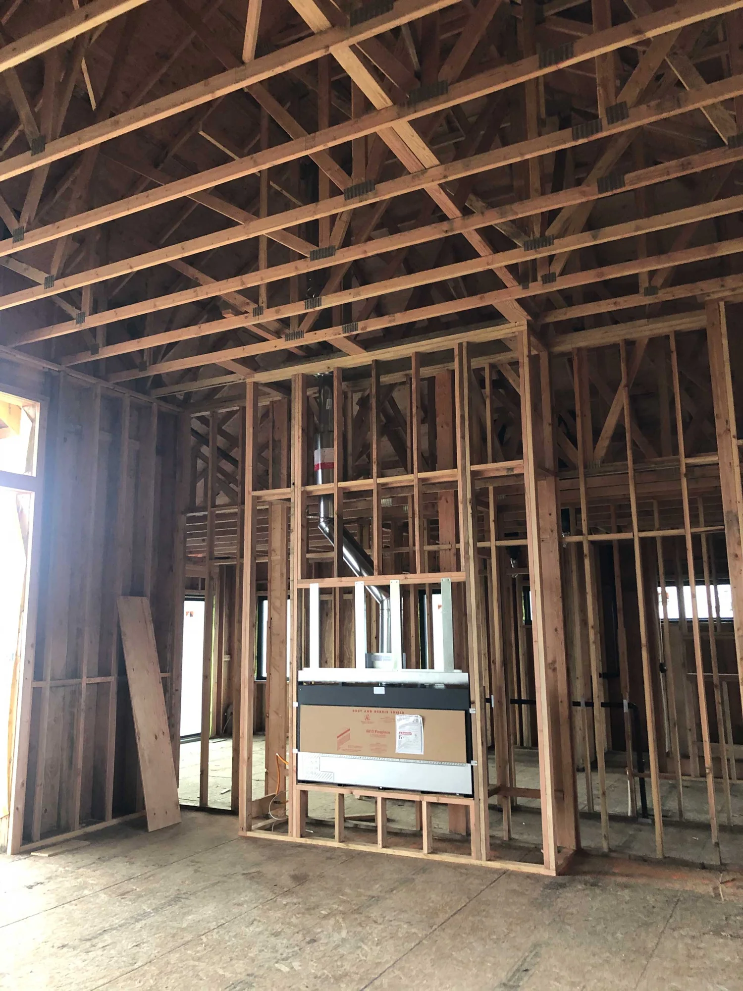 House interior under construction showing exposed wooden studs and ceiling trusses, a metal vent pipe and a boxed-in appliance rough-in at center.