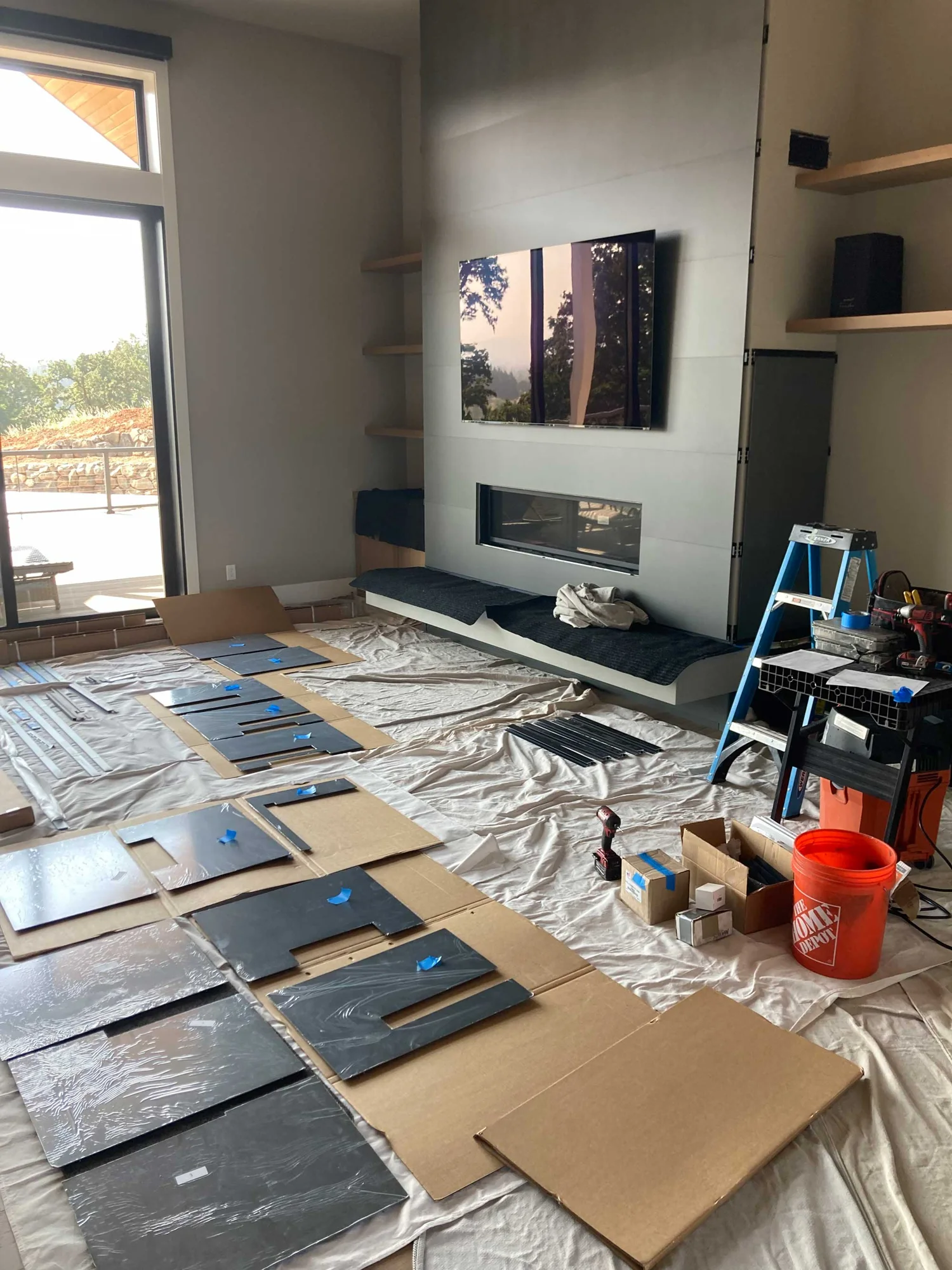Living room under renovation with TV mounted above fireplace, shelves, panels and cardboard on the floor, ladder, tools and an orange bucket.
