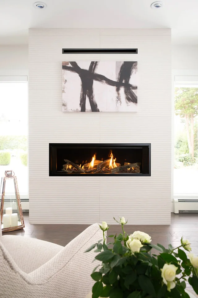 Modern living room with linear gas fireplace in a white textured wall, abstract black and white painting above, flames visible, light armchair and white roses in foreground