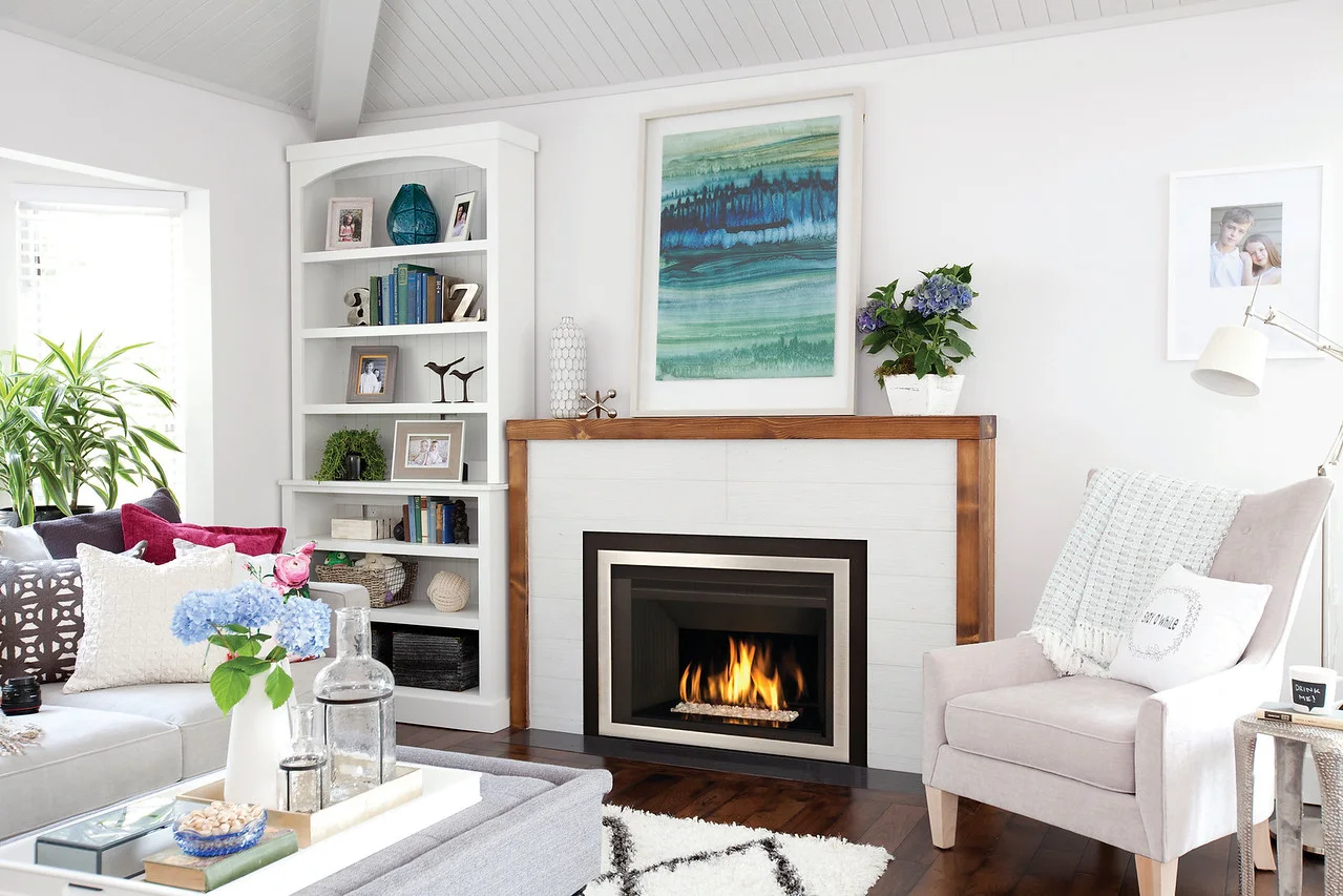 Bright living room with a lit modern fireplace with wooden mantel, white built-in bookshelf, upholstered armchair with throw, sofa, coffee table with vase of flowers