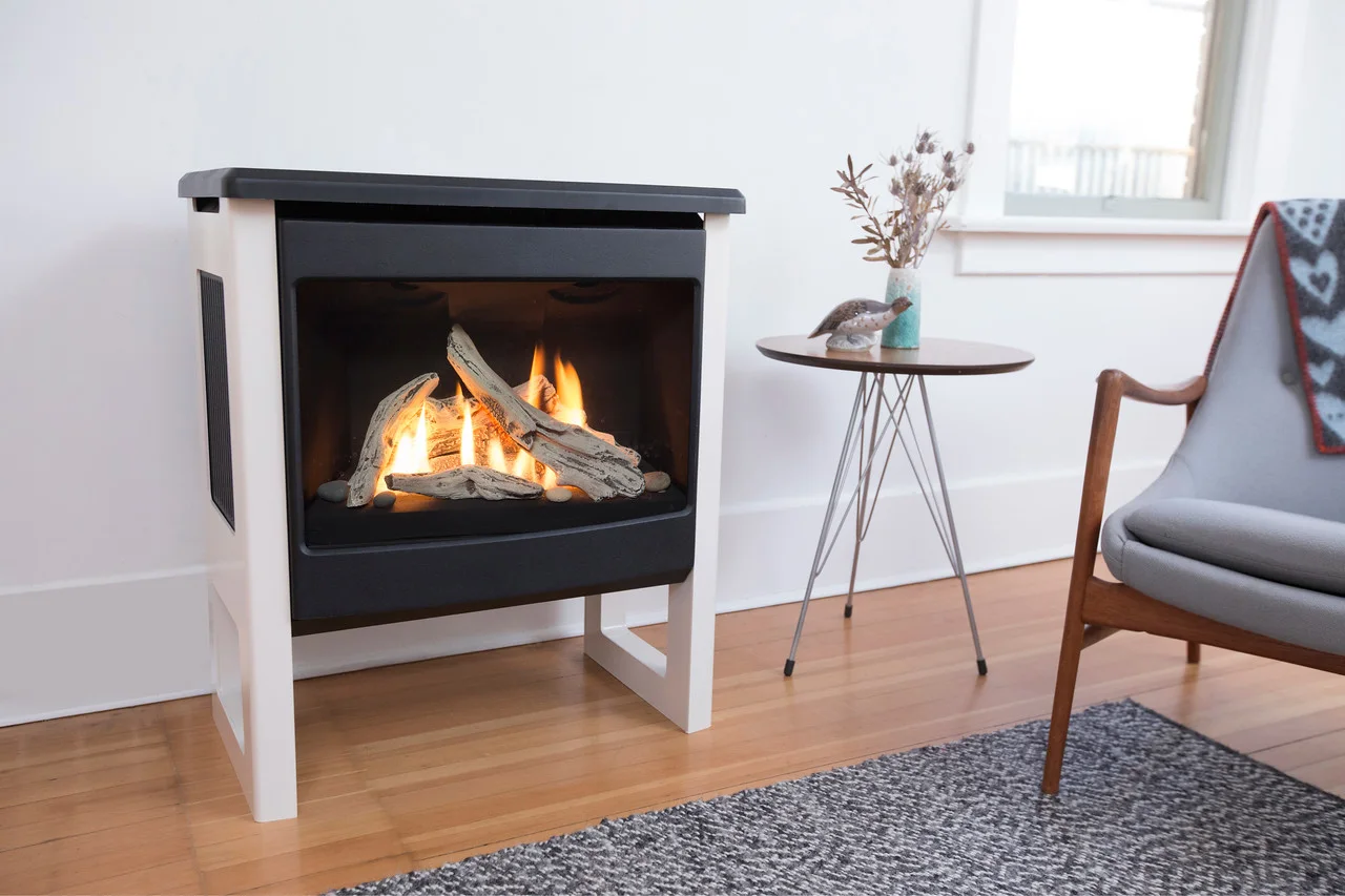 A modern, gas-burning fireplace next to a small round table with dried flowers and a bird statue. Nearby is a mid-century armchair with a blanket draped over its arm.