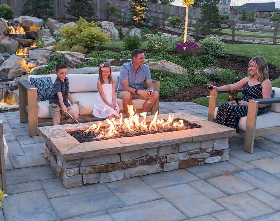 Family sitting around a stone fire pit on a backyard patio, kids roasting marshmallows near a landscaped waterfall.