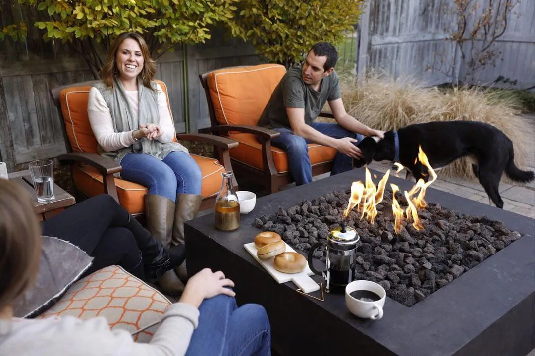 Three people seated on orange patio chairs around a lit outdoor fire pit with coffee, bagels, and a black dog