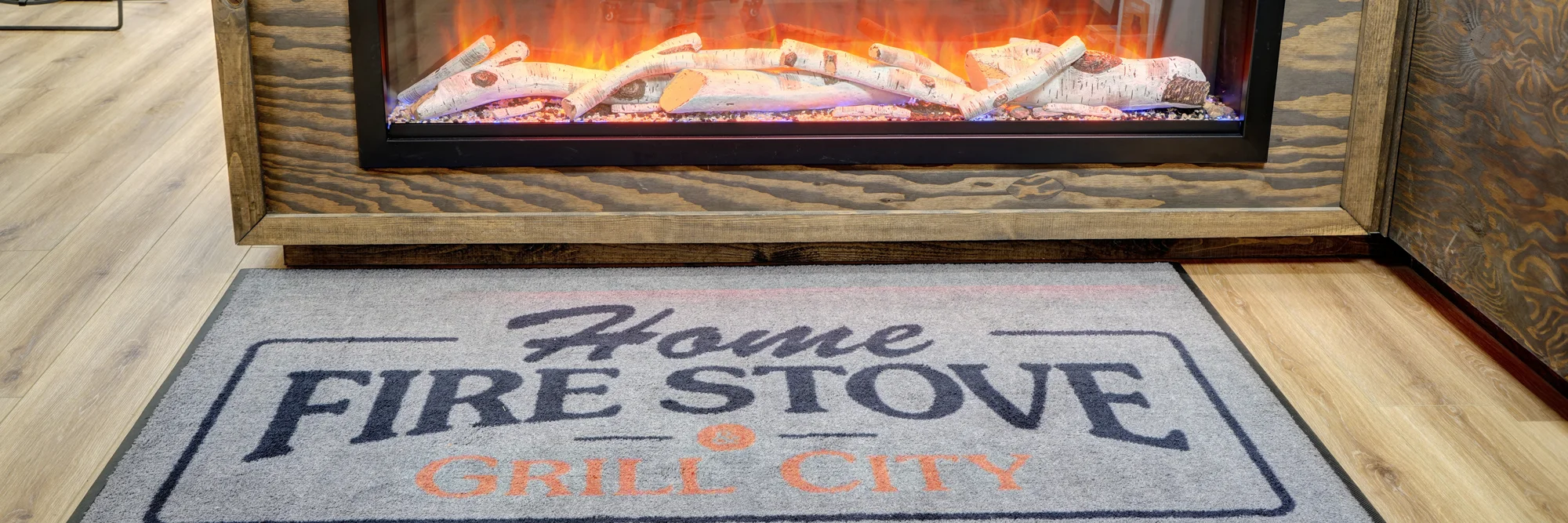 Electric fireplace with glowing logs and flames above a mat reading Home Fire Stove Grill City on a wood floor.