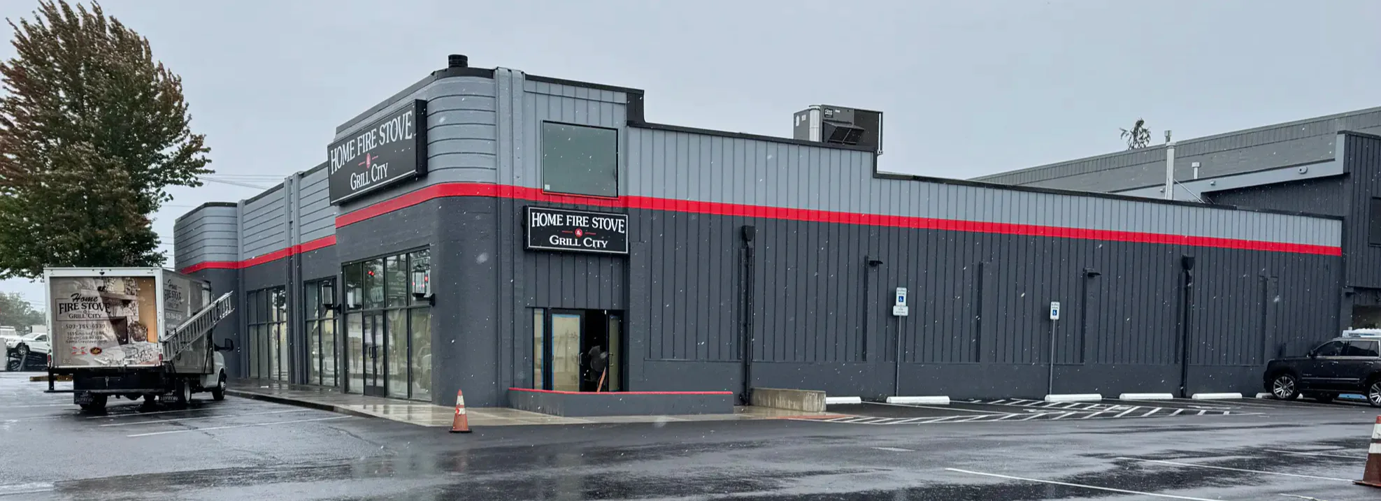 Gray commercial building with red stripe and Home Fire Stove Grill City sign on a wet parking lot