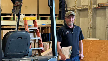 Two men standing beside an orange Doosan forklift in a warehouse with stacked boxes, ladder and plywood panels