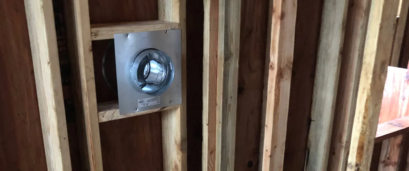 Partially constructed interior wall frame with wooden studs, showing an installed vent or pipe outlet with a metal flange in one of the stud bays. The floor is unfinished.