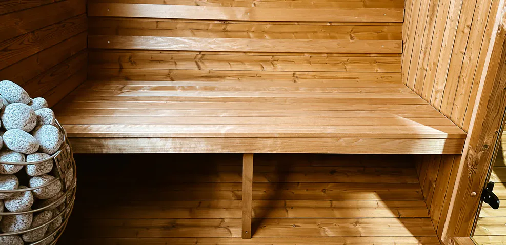 A wooden sauna with a bench, wooden paneled walls, and a basket of sauna stones. Sunlight filters in through a window, illuminating the interior.