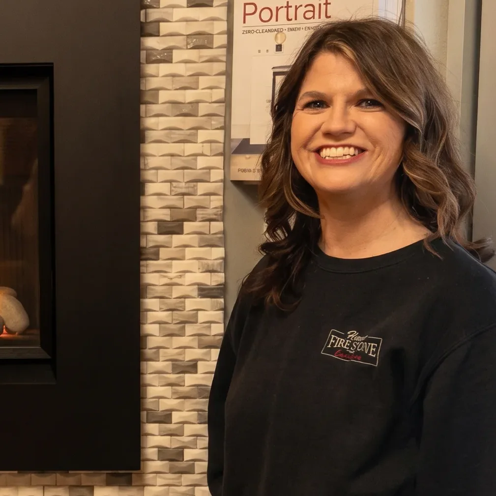 Smiling woman standing in a showroom beside a modern gas fireplace with round stones and textured tile wall