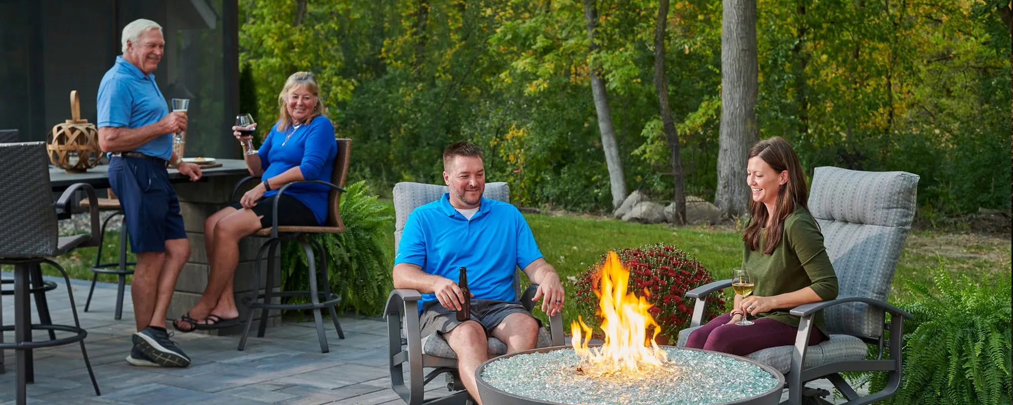 Four adults relaxing around a backyard fire pit on a stone patio, holding drinks and chatting