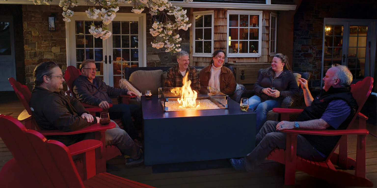 Six people sitting around an outdoor fire pit at night, engaged in conversation and laughter. They are seated on red chairs and a couch with drinks on the table and in hand. A house is in the background.