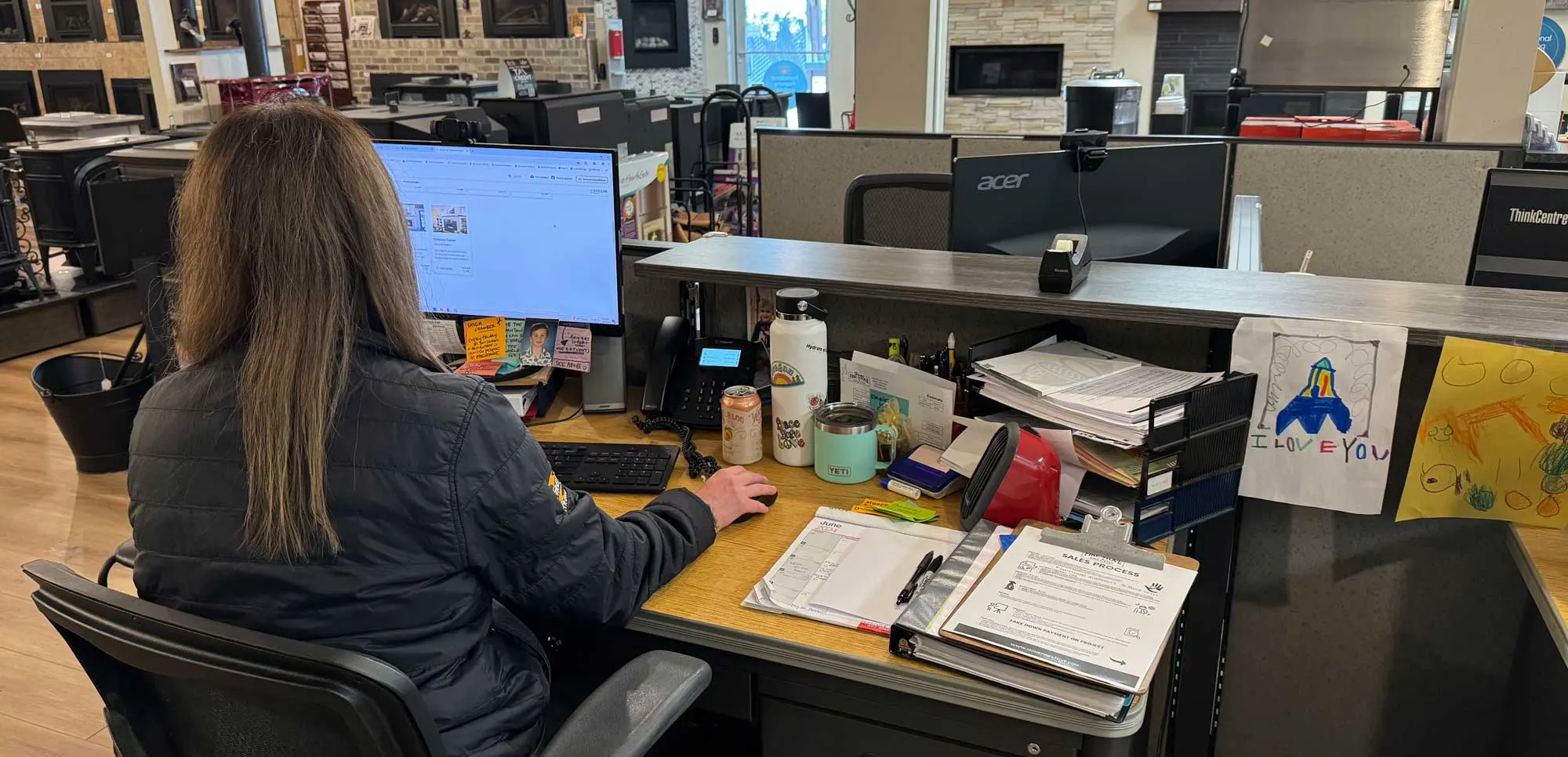 A person in a dark jacket works at a cluttered desk with a computer, phone, various papers, and children's drawings. The office has displays and bright lighting in the background.