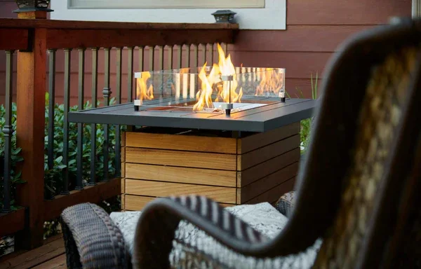 A wicker chair placed near a modern outdoor gas fire-pit with glass barrier