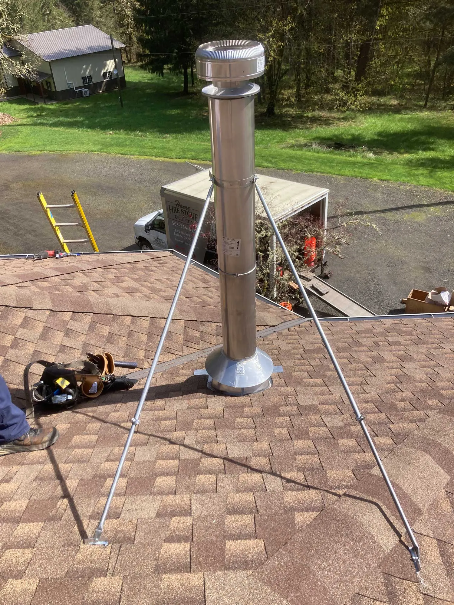 A new metal chimney pipe with support braces installed on a shingled roof. A person in work boots and a tool belt, a parked truck, and a ladder are also visible. A grassy yard is in the background.
