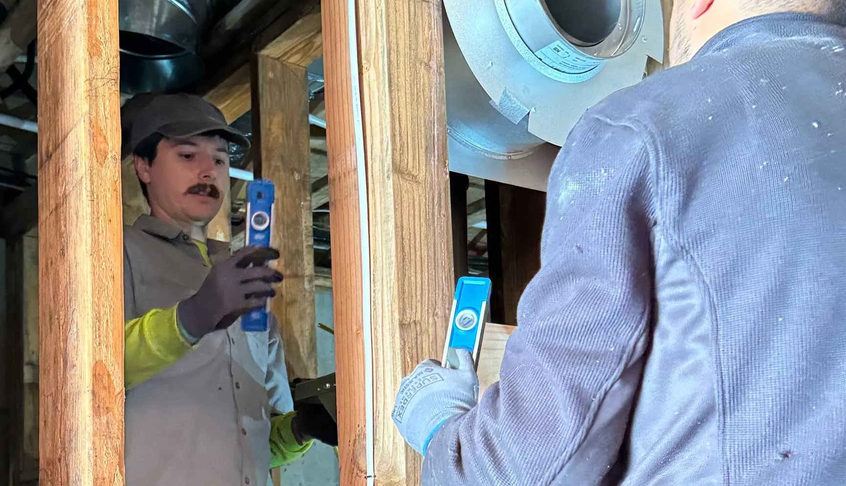 Two construction workers in a wooden frame structure, both holding blue levels. The worker on the left wears a grey shirt and cap while the worker on the right wears gloves and a blue jacket.