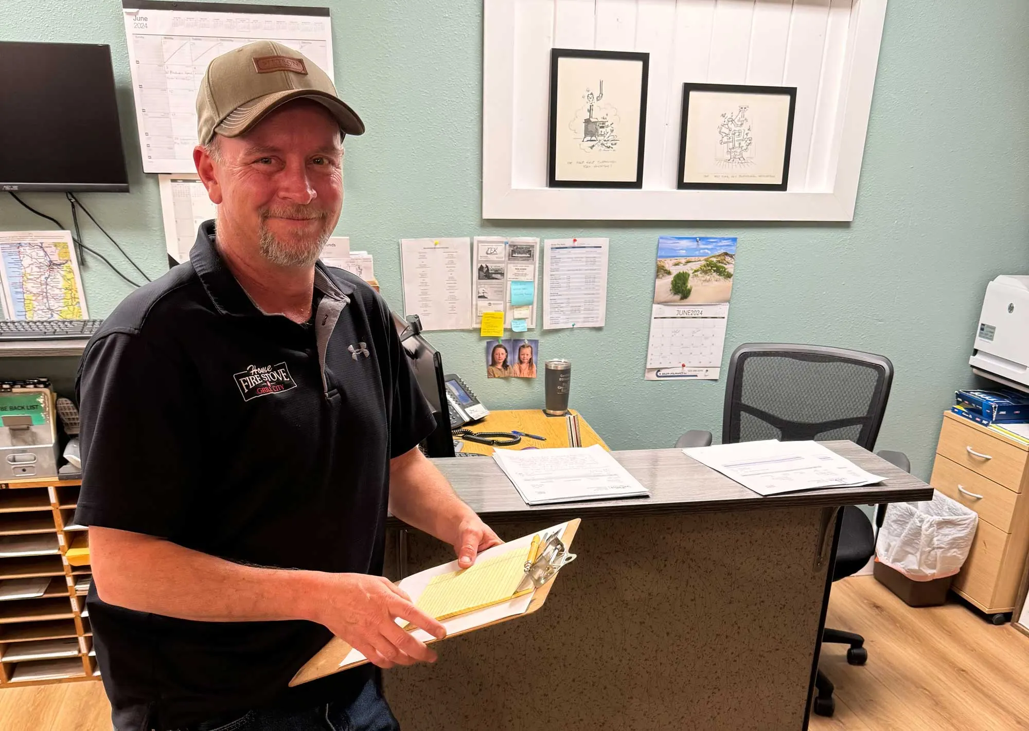 Man in a black shirt with a logo and a tan cap holds a clipboard, smiling in an office with a desk, chair, computer, wall calendar, and framed drawings on a green wall.