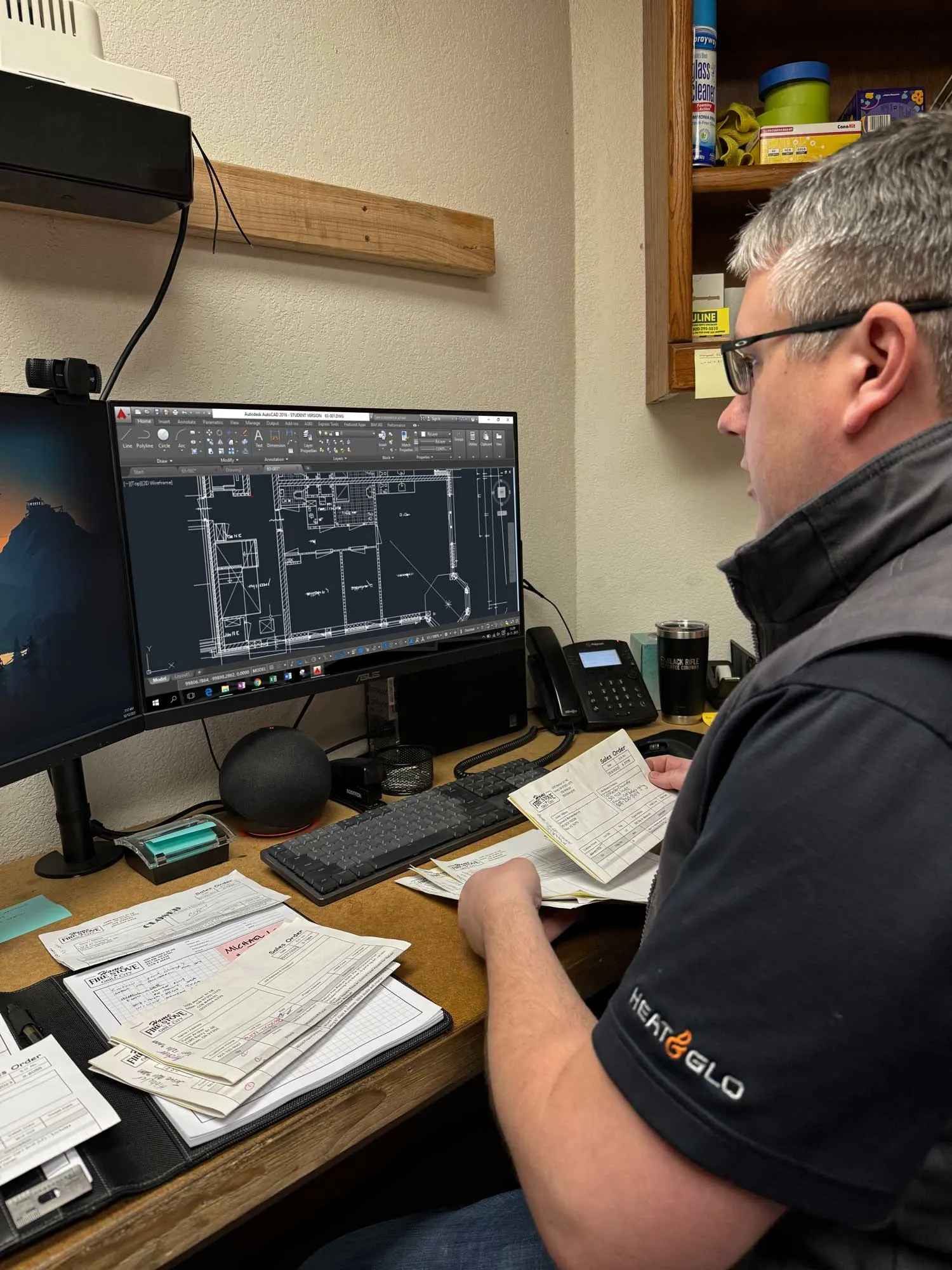 A man with glasses sits at a desk analyzing papers, while a computer monitor displays a digital floor plan on his screen. Office supplies are seen on the desk and shelf.