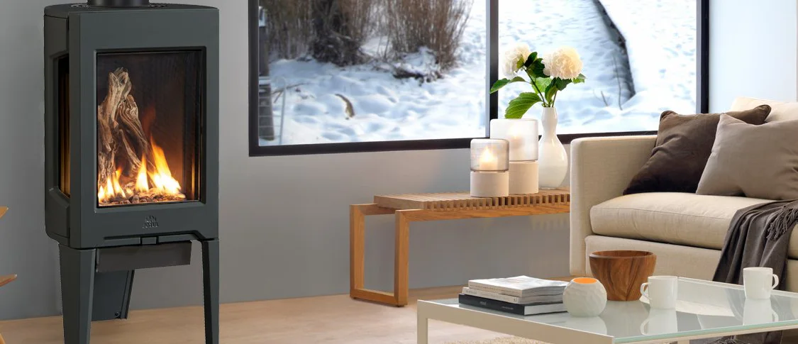 Modern living room with cozy fireplace, white couch with brown and gray pillows, wooden coffee table with books and candle, large windows showcasing a snowy outdoor scene.