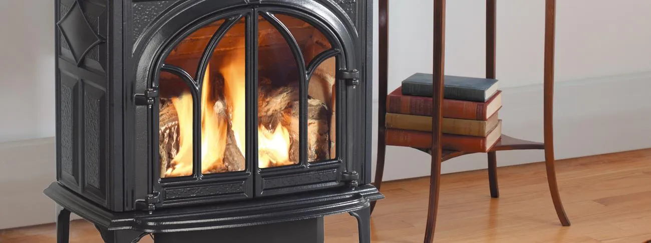 A black, ornate wood stove with a fire burning inside, sitting on a wooden floor next to a small side table with four stacked books on the lower shelf.