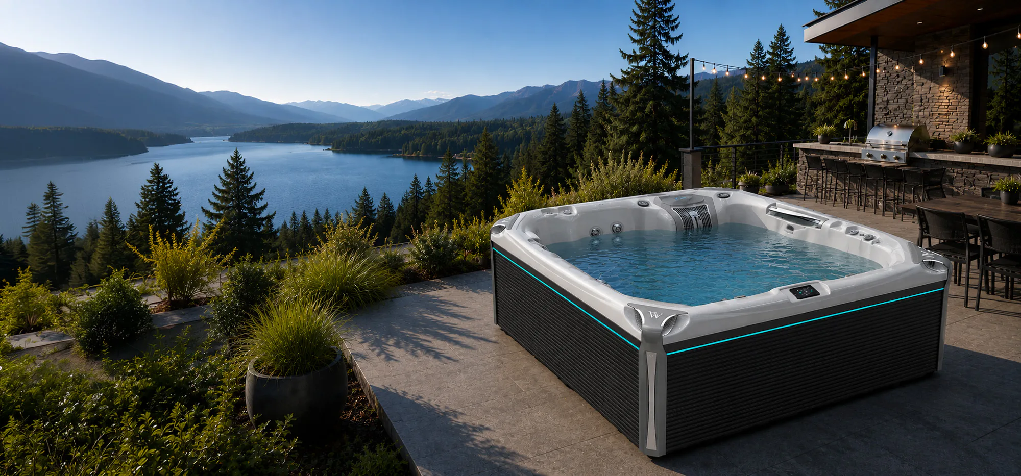 Patio hot tub filled with water overlooking forested lake and distant mountains, outdoor kitchen and string lights.