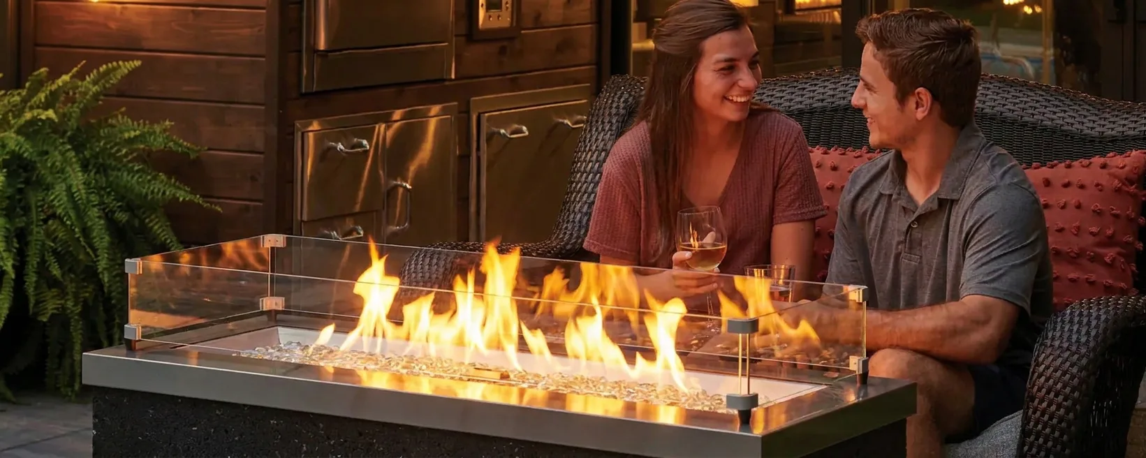 Young couple sitting on wicker patio chairs by a modern rectangular fire pit with glass wind guard, smiling and holding drinks in a warm evening outdoor setting.