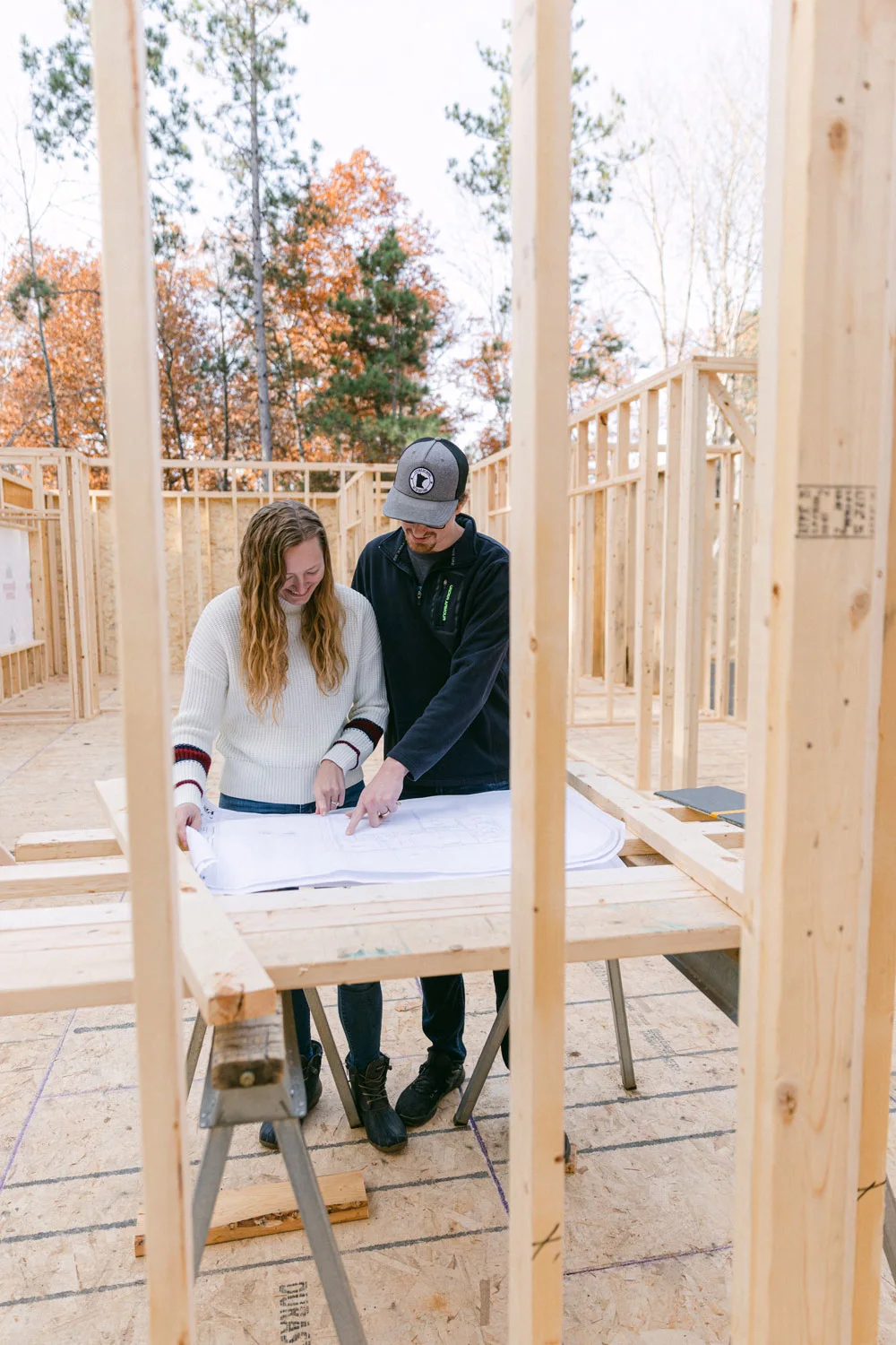 Two people standing inside a partially built house, looking at architectural blueprints on a wooden table. The framework of the house and fall trees are visible in the background.