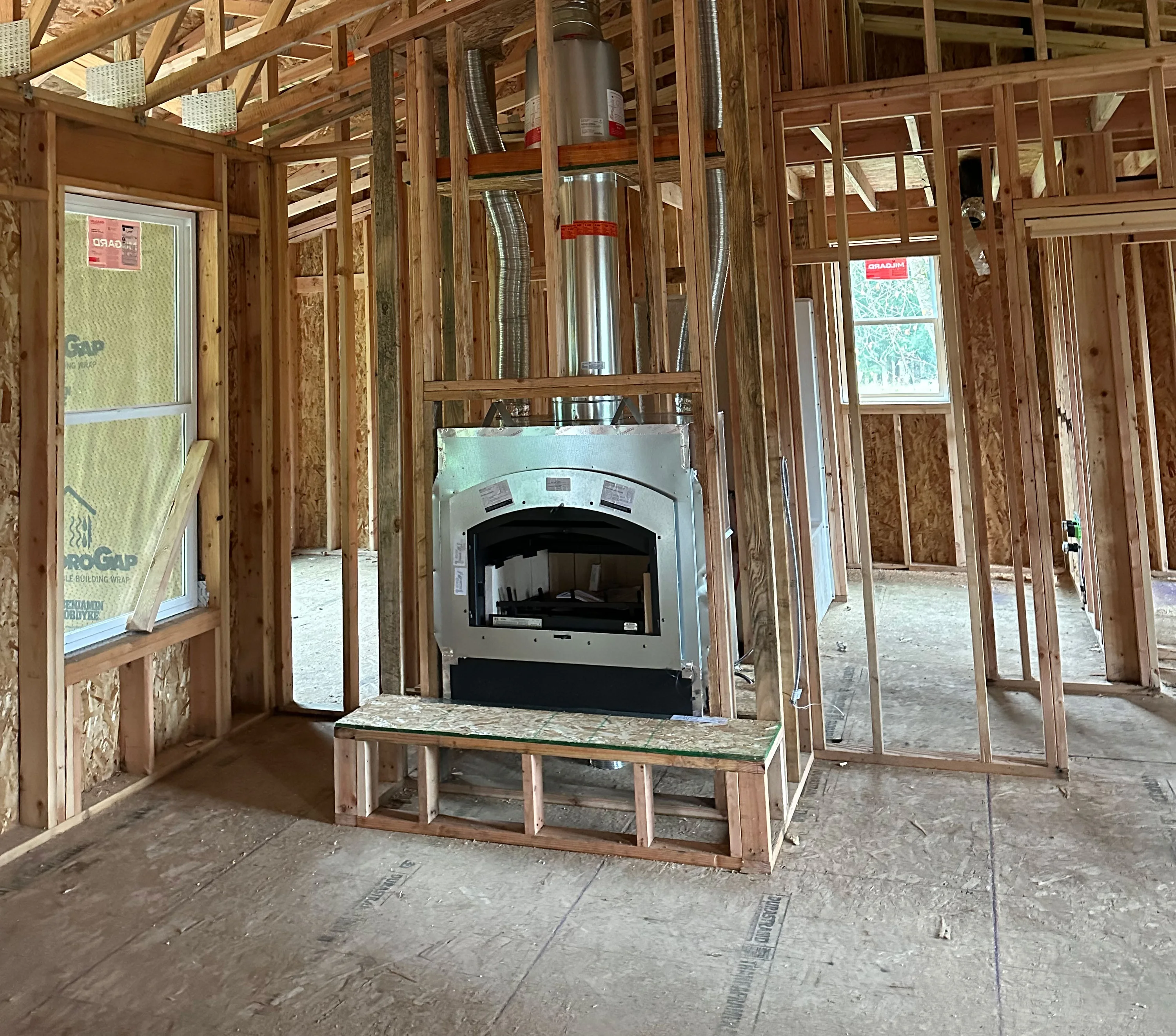 A partially constructed home with exposed wooden framing, an installed metal fireplace, and insulation visible through a window on the left. Sunny background outside window.