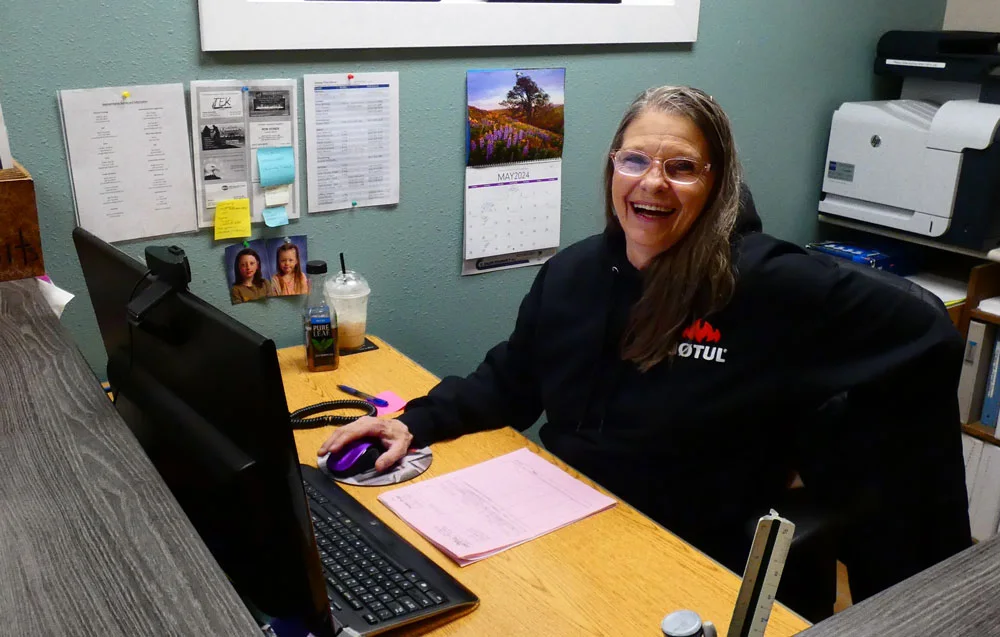 A woman wearing glasses and a black hoodie is sitting at a desk, smiling at the camera. She is using a computer mouse with one hand, and there are documents, a beverage, and family photos on the desk.