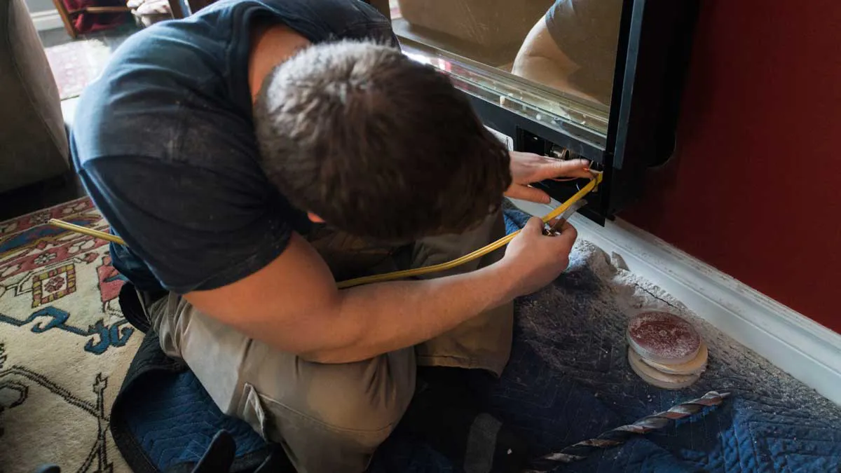 Man crouched by a fireplace installs wiring, surrounded by dust and tools, including a drill bit and tape. A colorful patterned rug is partially visible on the floor.
