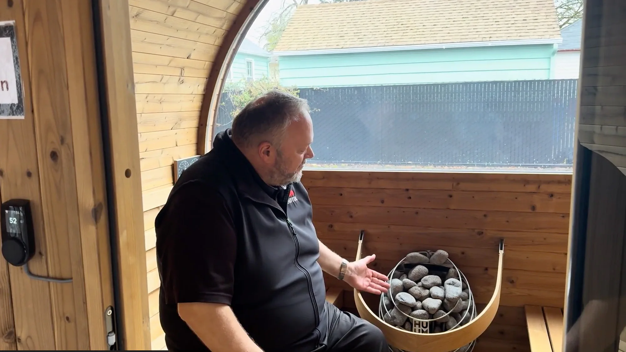 Man seated inside a wooden sauna, gesturing toward a basket of sauna stones by a round window