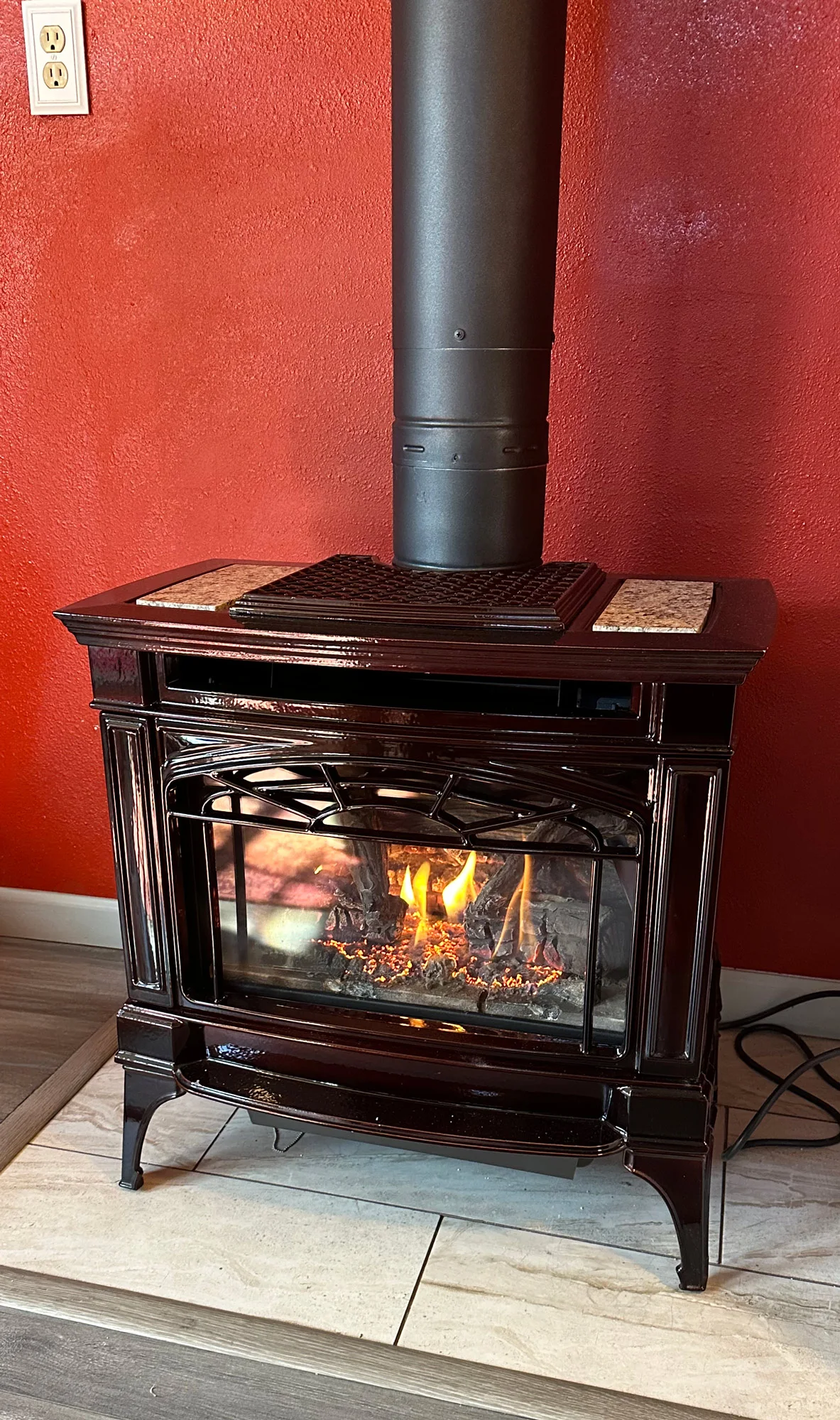 A black stove with a fire burning inside sits against a textured red wall next to an electrical outlet. The stove is on a tiled base with a wooden floor surrounding it.