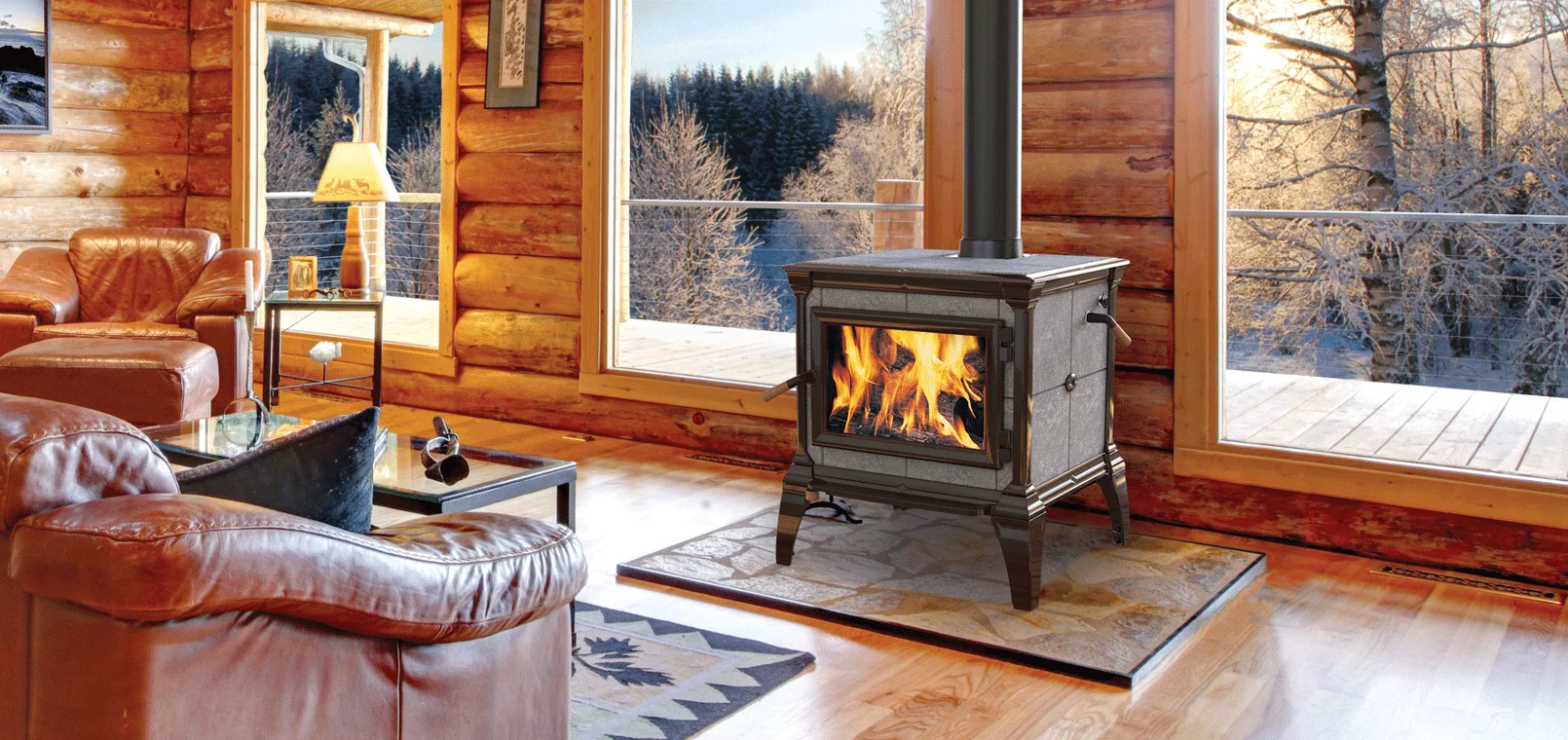 A cozy room with a wood-burning stove, large windows with a snowy forest view, and leather chairs. A lamp, side table, and a patterned rug add to the warm ambiance.